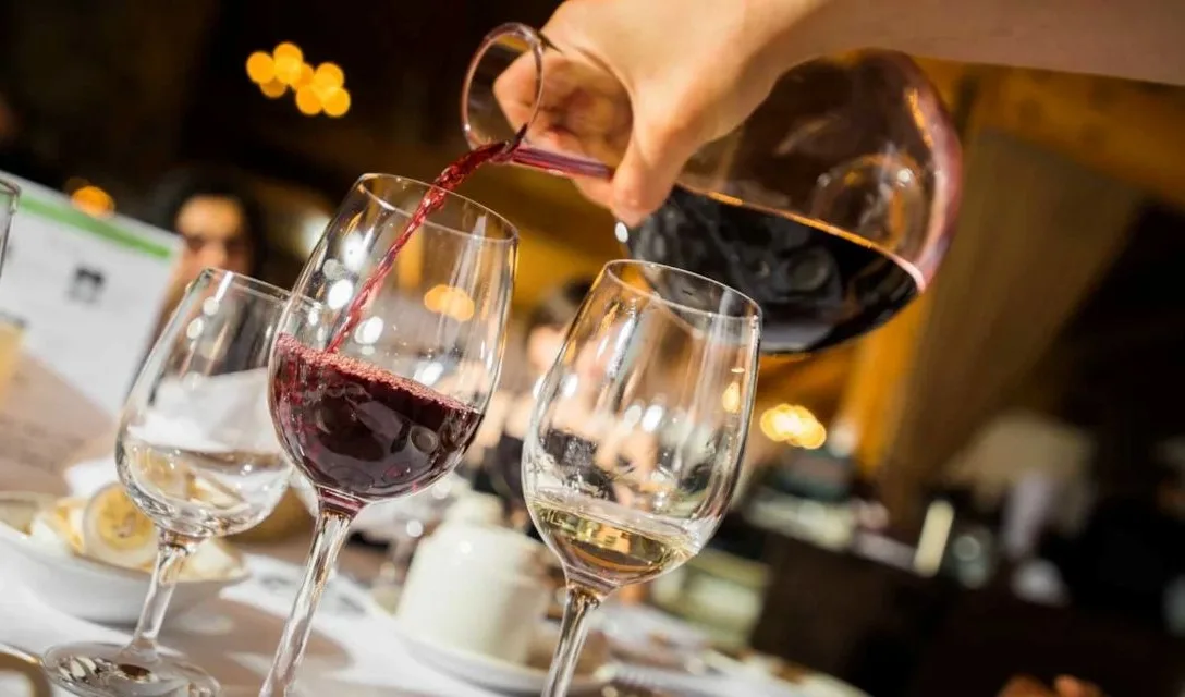 A hand is pouring a glass decanted full of red wine into one wine glass. The glass to the right of this has red wine filling it a fifth of the way up.