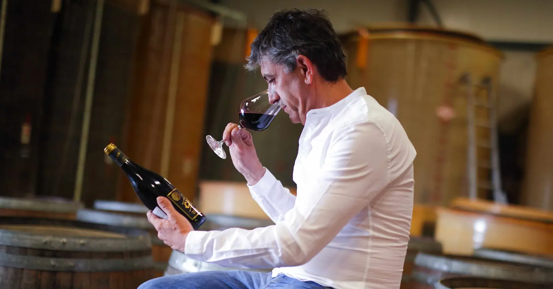 Winemaker Herve Sabardeil in a white shirt sitting on a barrel holding a bottle of Cabalié Originé wine and sipping from a glass.