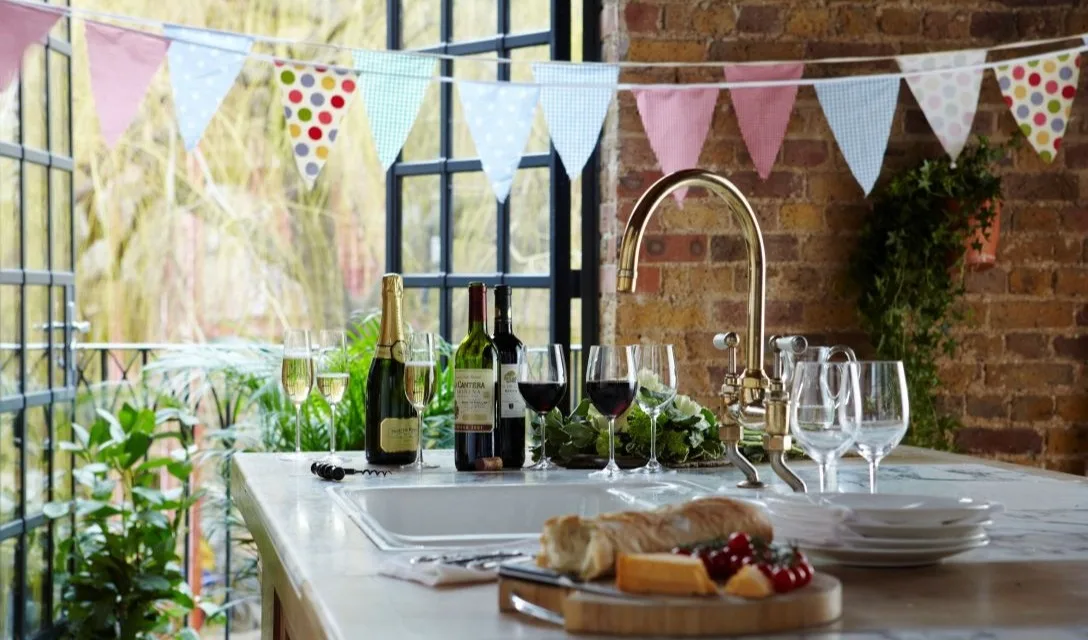 Wine bottles and glasses arranged on a kitchen island beside a sink, with large windows and bunting decorations overhead.