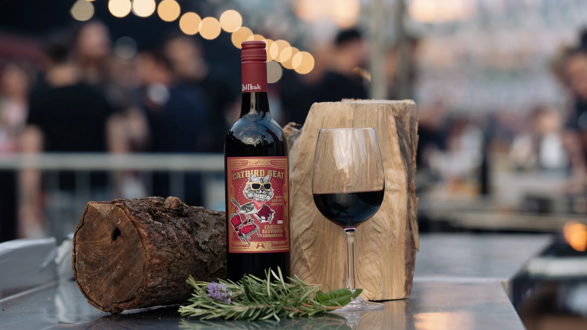 Bottle of Catbird Seat red wine beside a filled wine glass, logs and rosemary on a metal table with blurred lights behind.