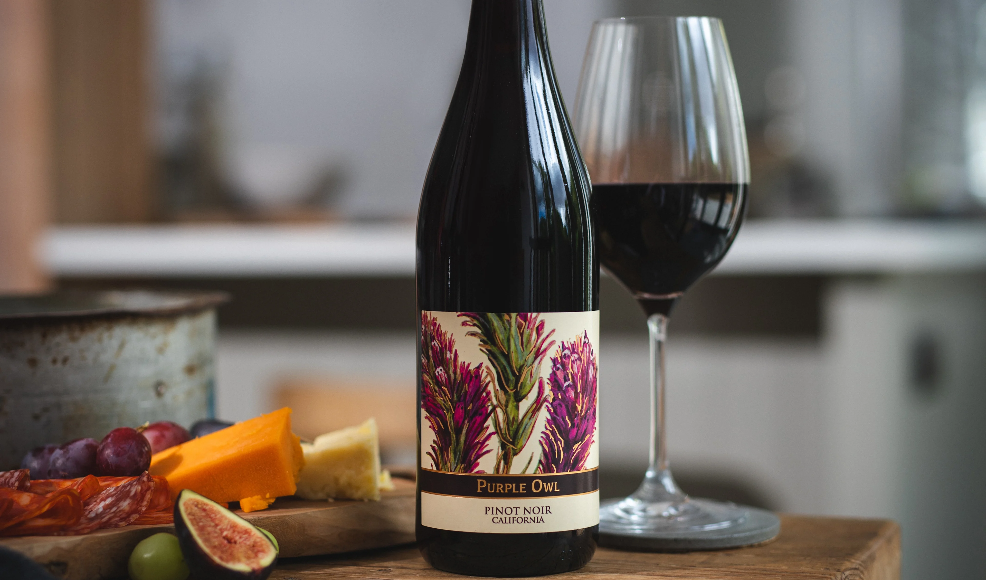 Bottle of Pinot Noir displayed beside fresh veg, figs and cherries on a wooden board against a dark backdrop.