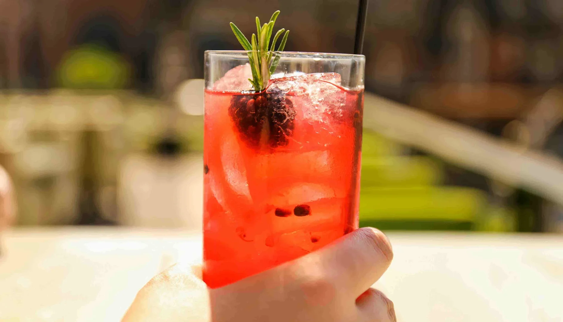 Hand holding a tall glass of pink mocktail with ice, blackberry, rosemary garnish, and a straw in an outdoor setting.