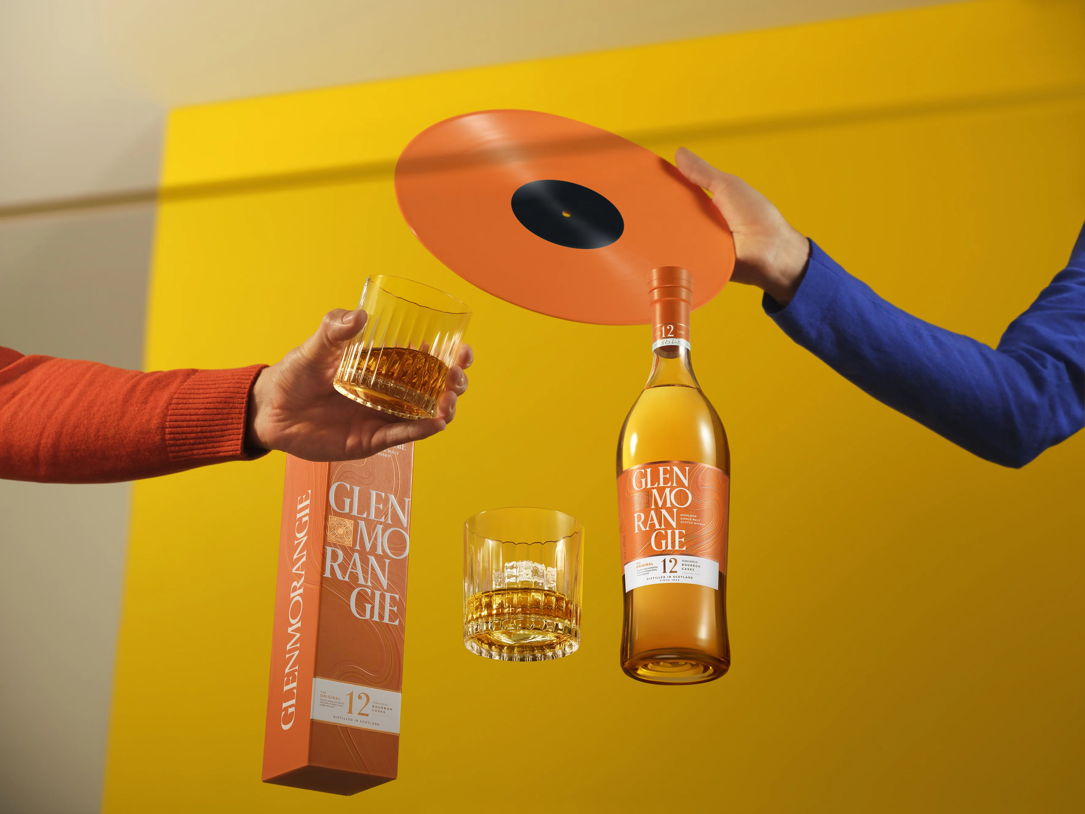A Glenmorangie whisky bottle, orange box, and two glasses with amber liquid, alongside an orange vinyl record, against a bright yellow background.