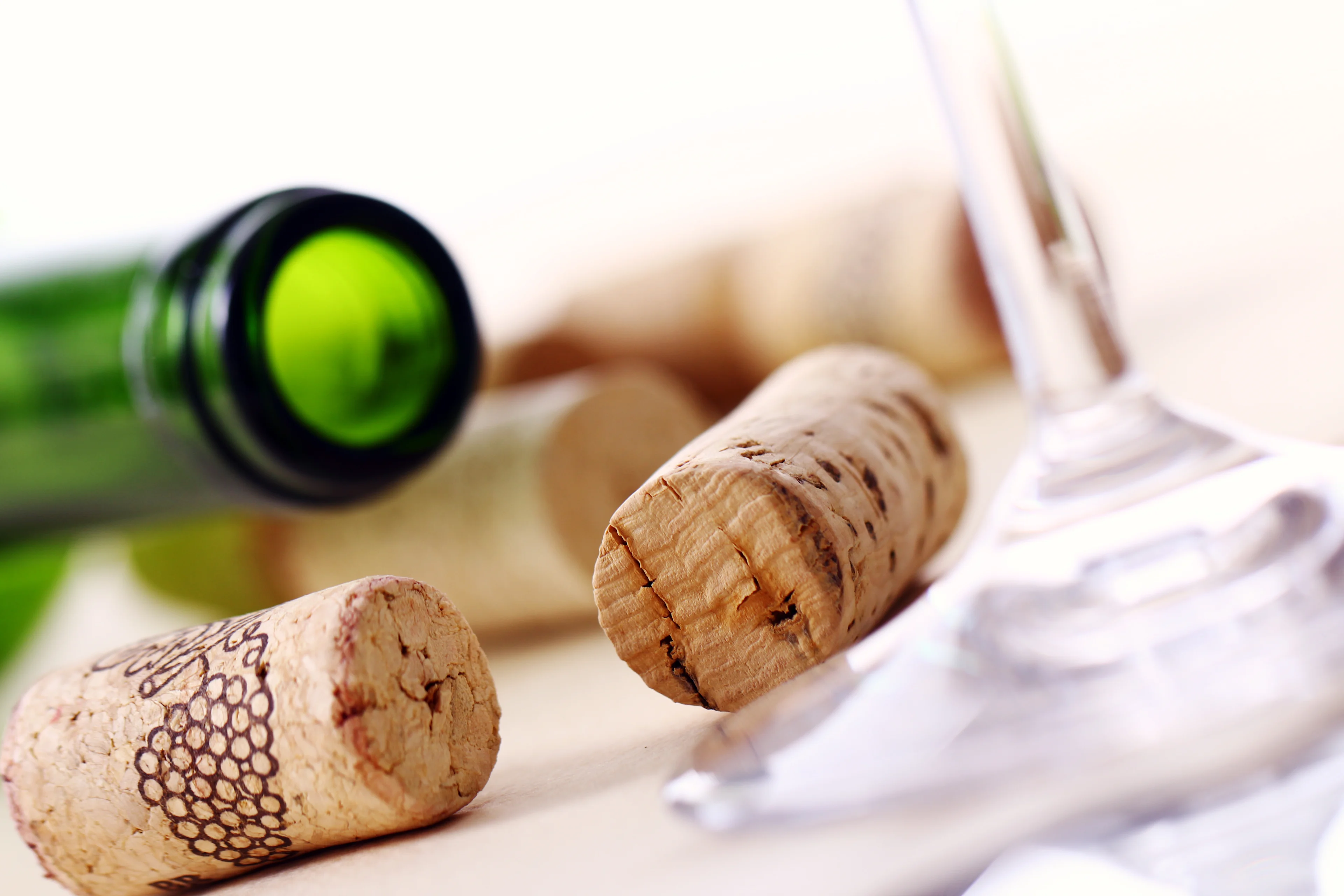 Several natural wine corks scattered on a light surface next to the base of a clear wine glass and the neck of a green glass wine bottle.