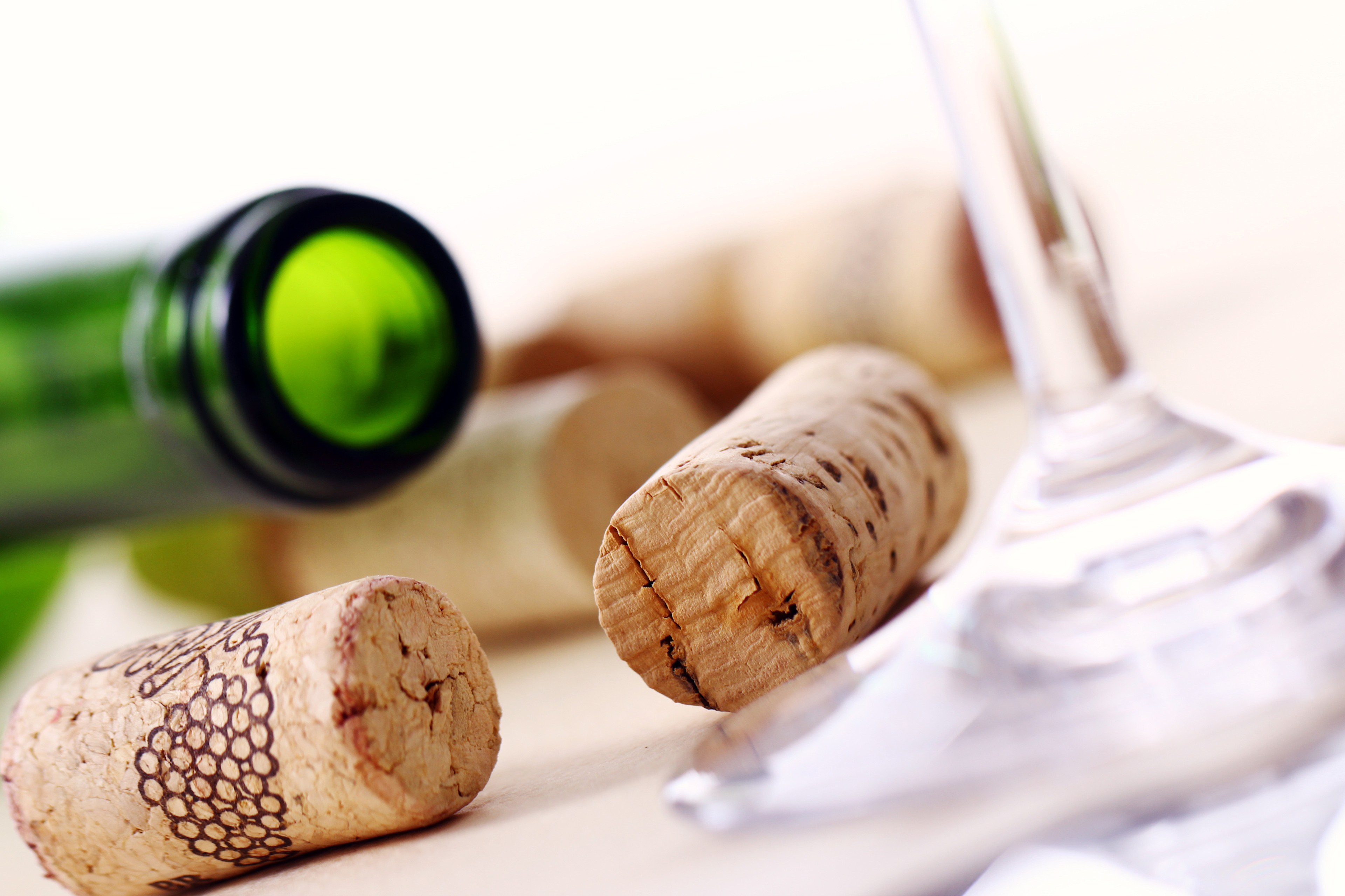 Several natural wine corks scattered on a light surface next to the base of a clear wine glass and the neck of a green glass wine bottle.
