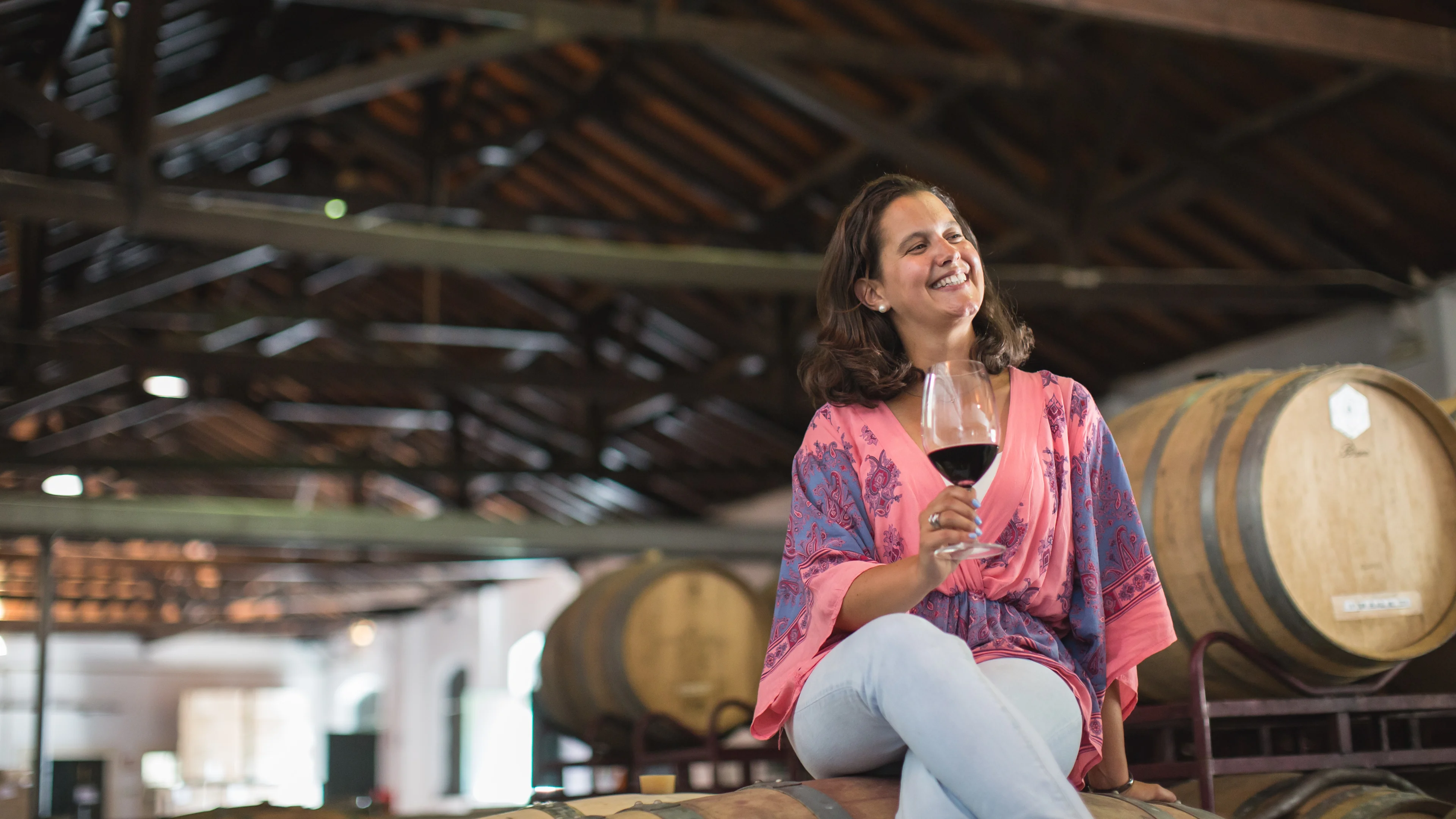 Person seated on a wine barrel holding a glass of red wine inside a winery with oak barrels in the background.