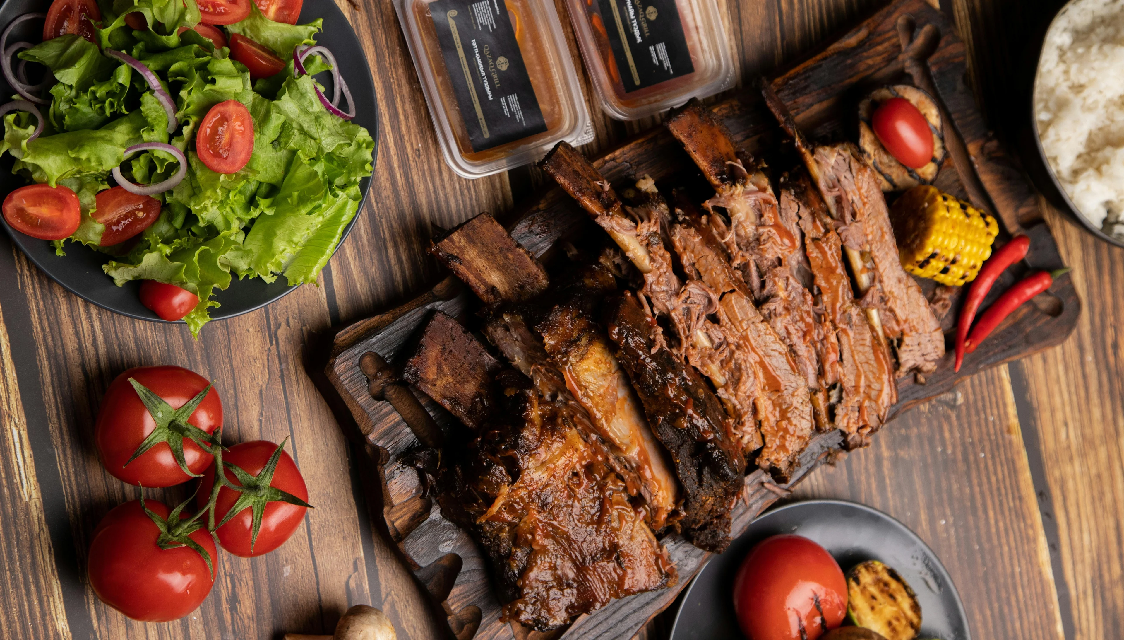 Barbecue ribs on a wooden board with sauce, corn, and chilies, served with a fresh salad, tomatoes, and rice on a rustic table.