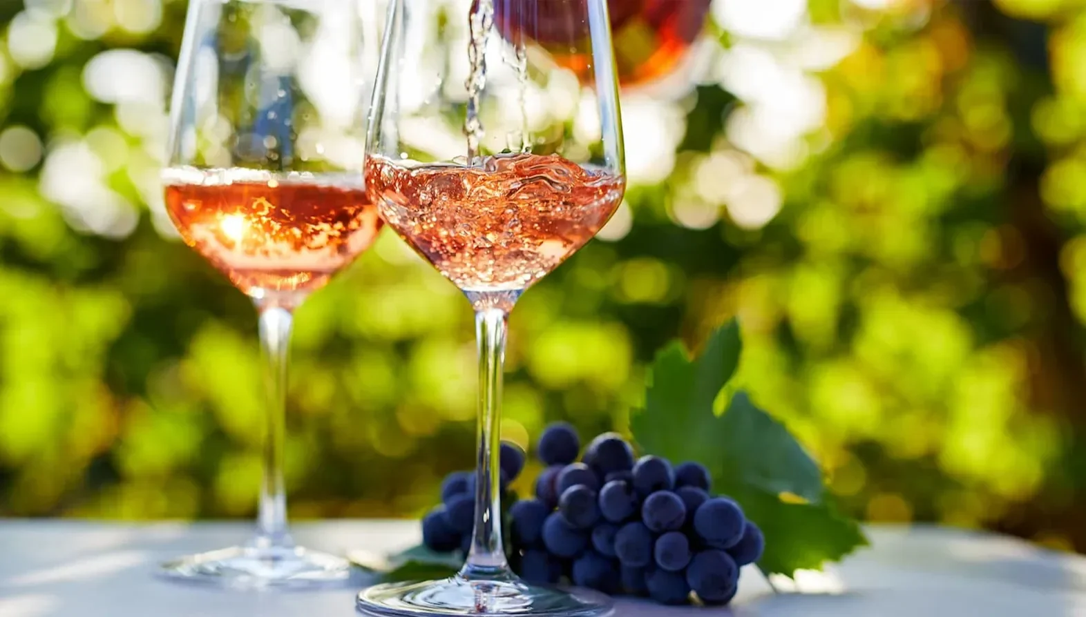 Two glasses of rosé wine being poured beside a small bunch of dark grapes outdoors.