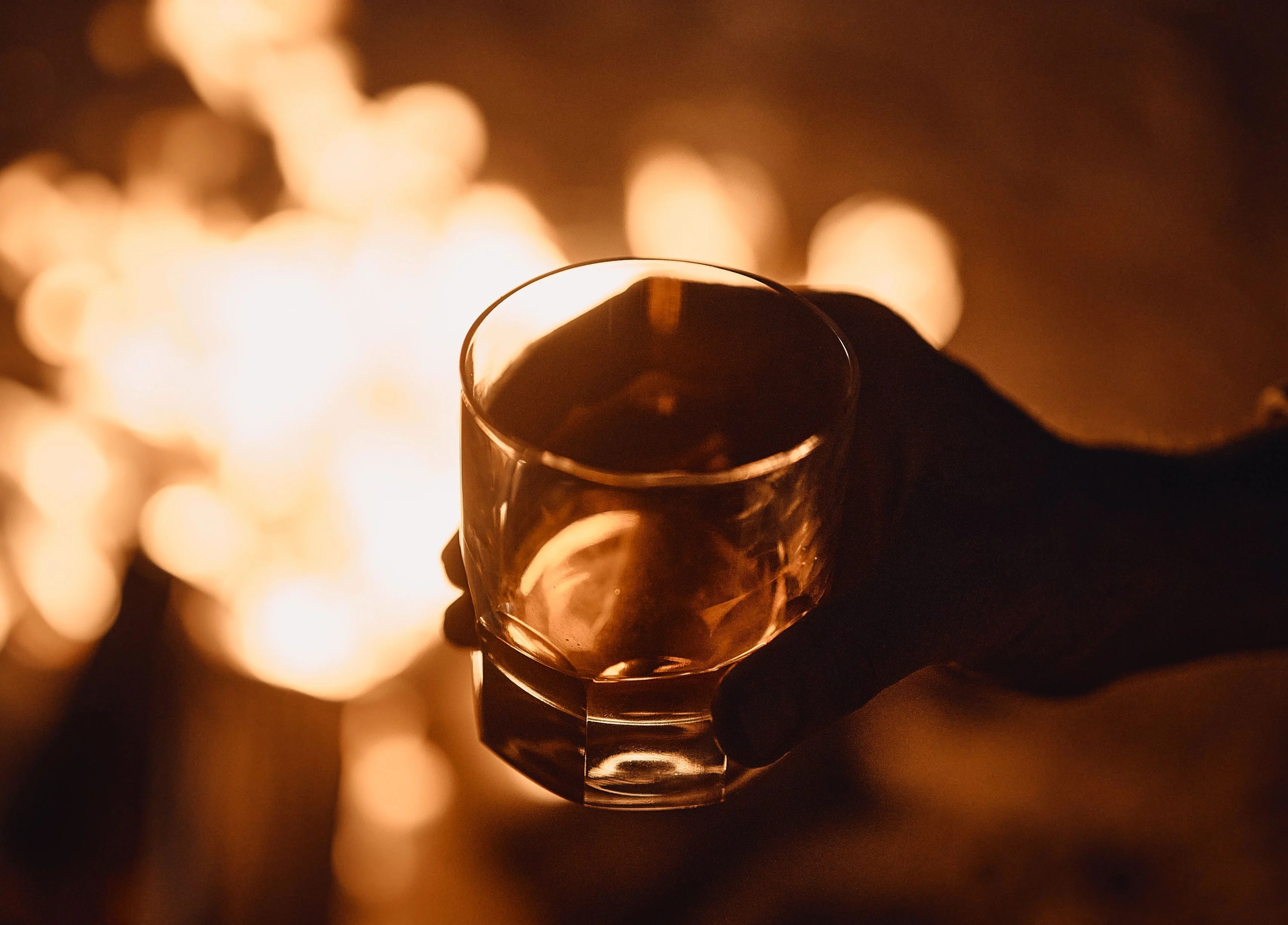 A hand holding a short glass of whisky with ice in front of a warm, blurred firelight background, creating a cozy and intimate atmosphere.