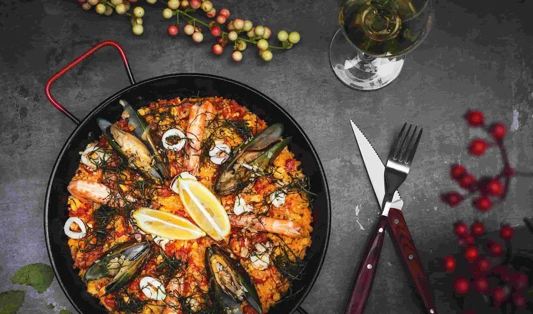 A full pan of paella set on a dark grey slate backdrop, with wooden-handled knife and fork, a glass of alcohol-free white wine and two lemon wedges on top of the paella for garnish.