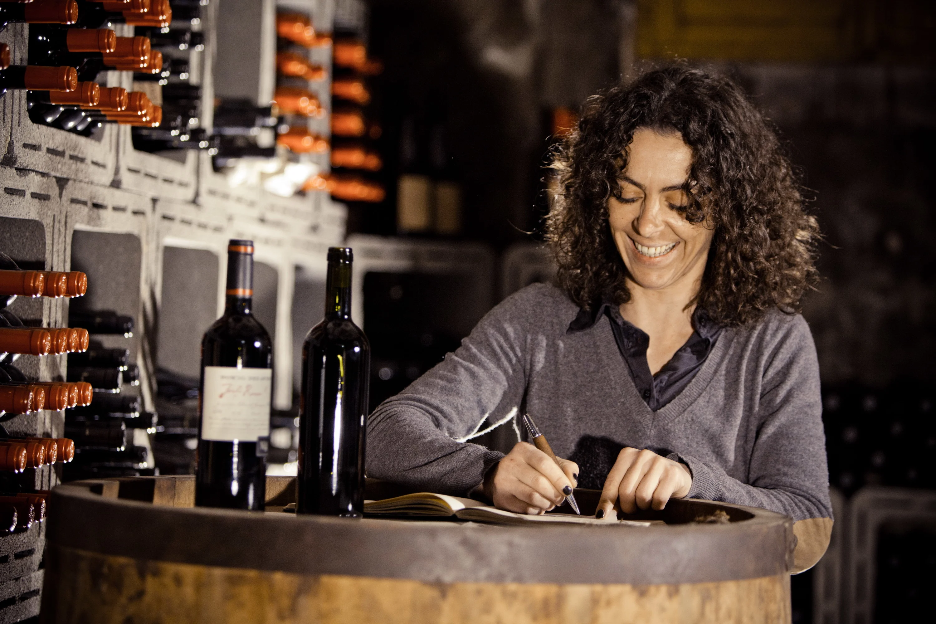 A female winemaker with dark curly hair sitting by a large barrel. There are wine bottles on the barrel and in the background. She is smiling and writing notes on a pad.