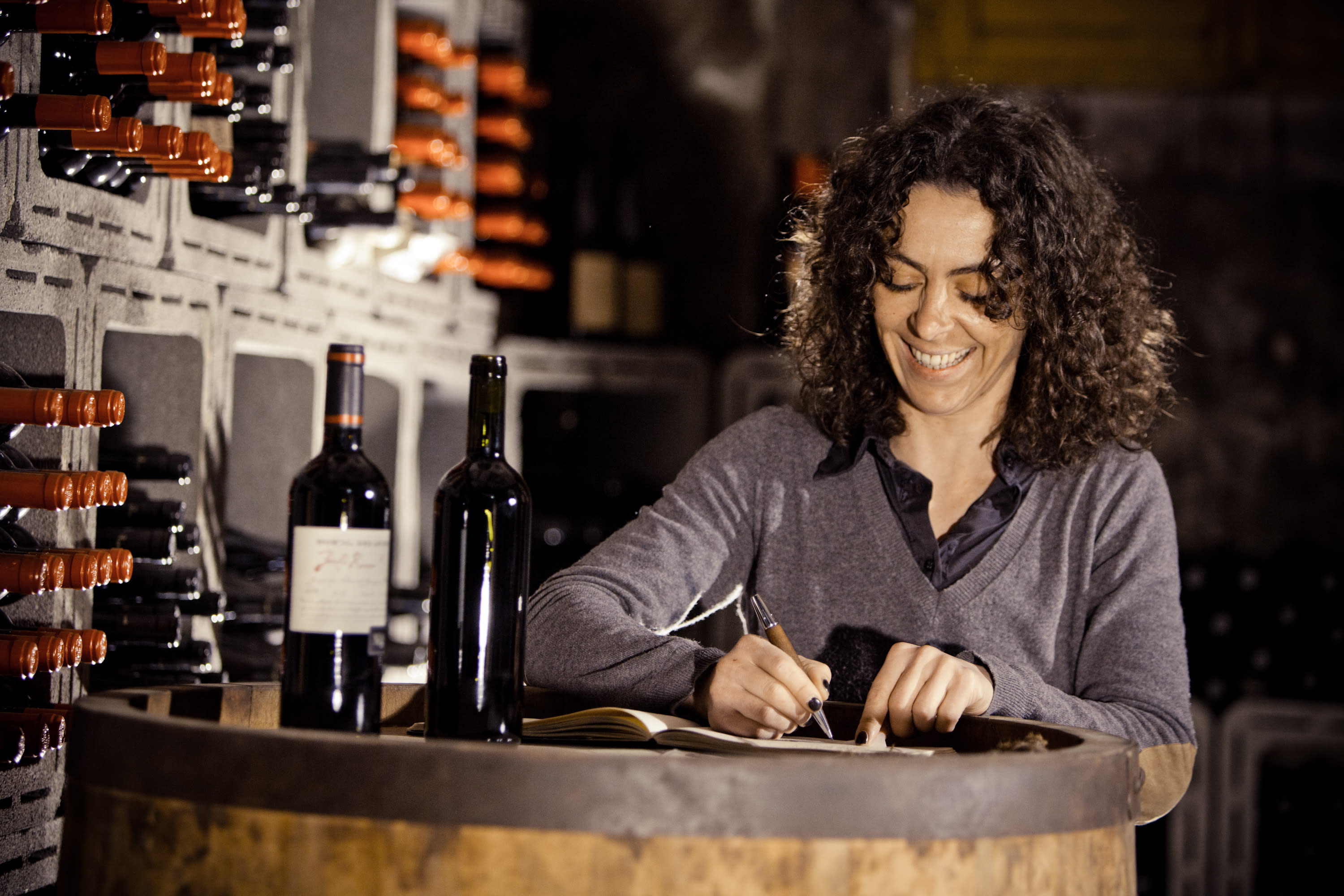 A female winemaker with dark curly hair sitting by a large barrel. There are wine bottles on the barrel and in the background. She is smiling and writing notes on a pad.