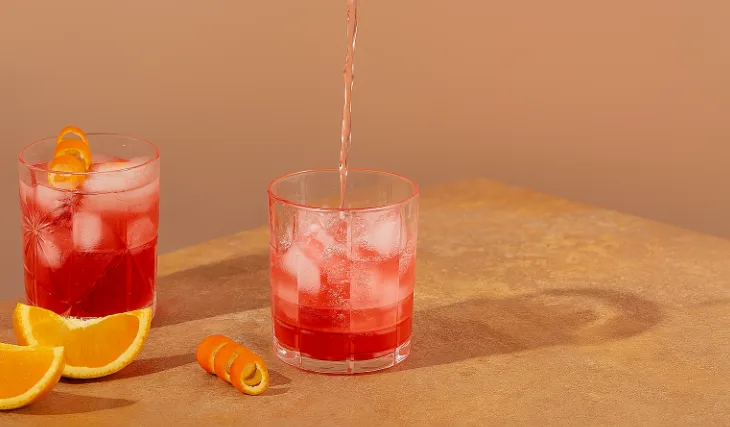 Two glasses of red cocktail with ice and orange peel garnish, one being filled, on a light brown surface with orange slices.