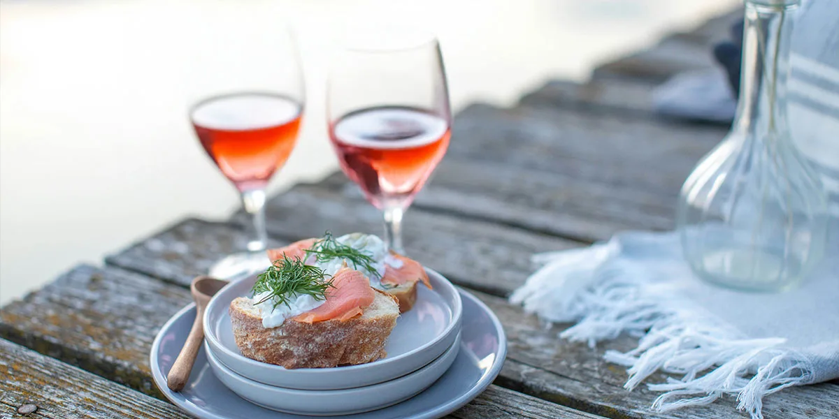 Two plates of smoked salmon on bread with cream and dill beside glasses of rosé by the water.
