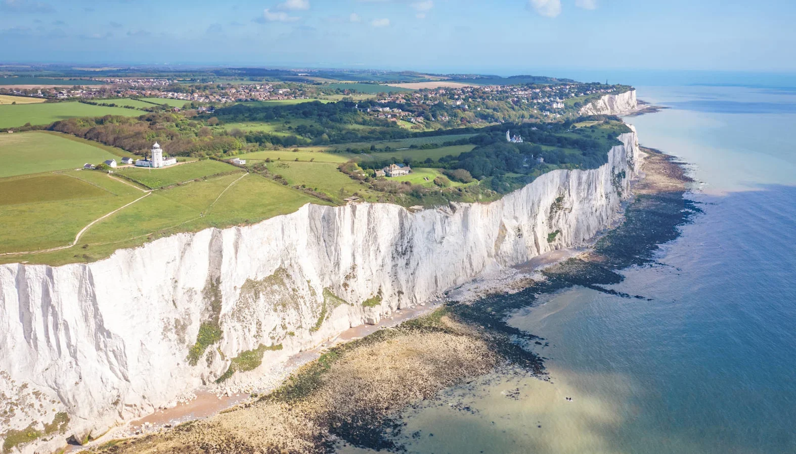 Aerial view of the White Cliffs of Dover with grassy fields, a lighthouse, and calm sea under a clear blue sky.