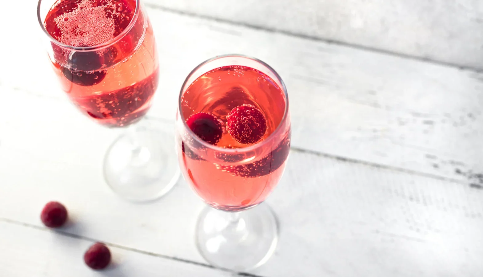 Two champagne flutes with pink sparkling cocktail and raspberries inside.