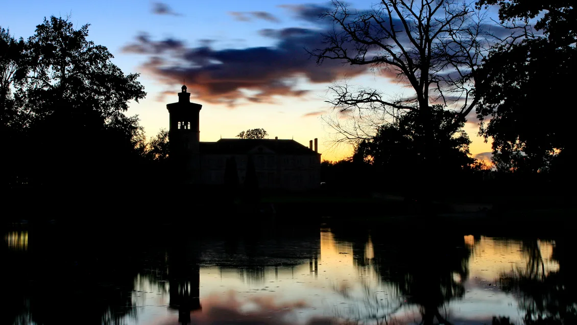 Silhouette of a château reflected in a still lake at dusk, framed by trees and twilight clouds.