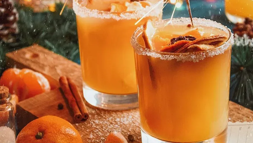 Two glasses of mulled drink with sugared rims, dried fruit garnish, and a lit sparkler, surrounded by oranges and cinnamon sticks.