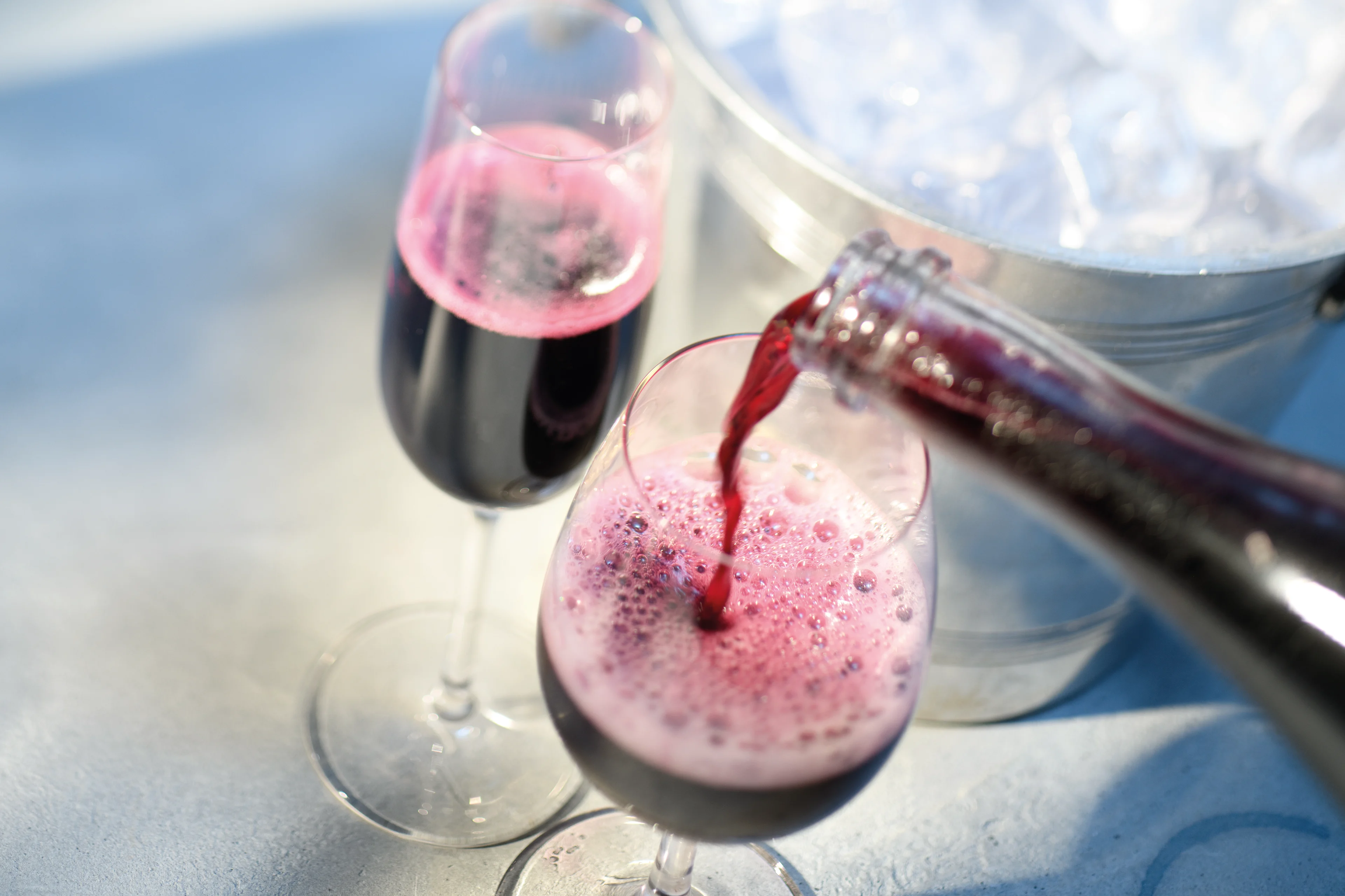 Red sparkling wine being poured into a flute glass, creating a frothy surface, with another filled glass nearby and a metal ice bucket in the background on a light-colored surface.