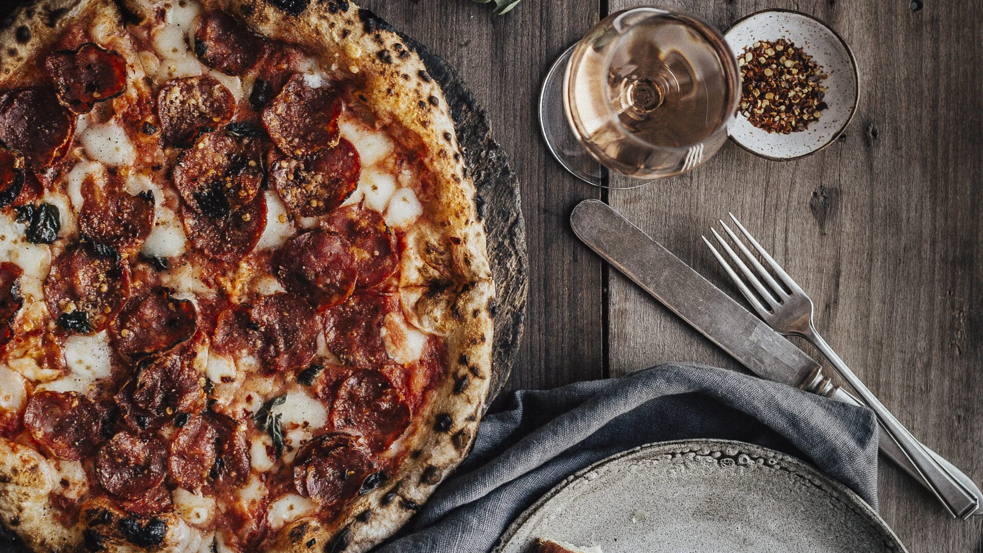 Pepperoni pizza on a wooden table beside a glass of rosé wine, cutlery and a dish of chilli flakes.