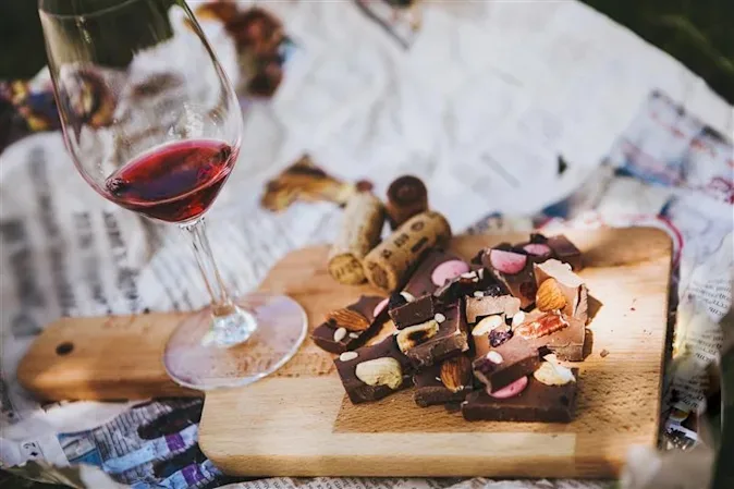 Glass of red wine and pieces of nut‑studded chocolate on a wooden board set on newspaper outdoors.