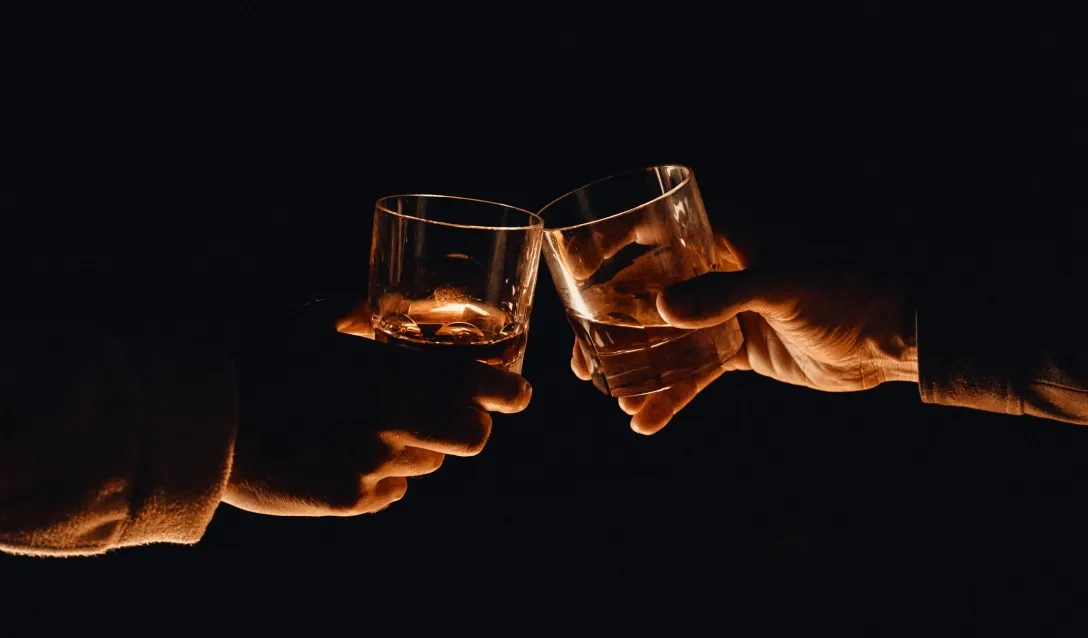Two hands clinking short glasses of whisky with ice against a dark background, illuminated by warm light highlighting the amber liquid.