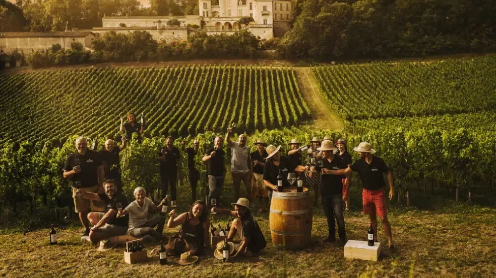 Group gathered among vineyard rows with wine bottles and barrels, overlooking a château at sunset.