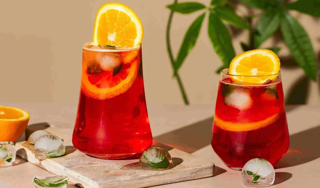Two cocktail glasses filled with alcohol-free red cocktails, garnished with a slice of orange and glass each, The glasses are set on a table with ice cubes nearby and half a cut orange.