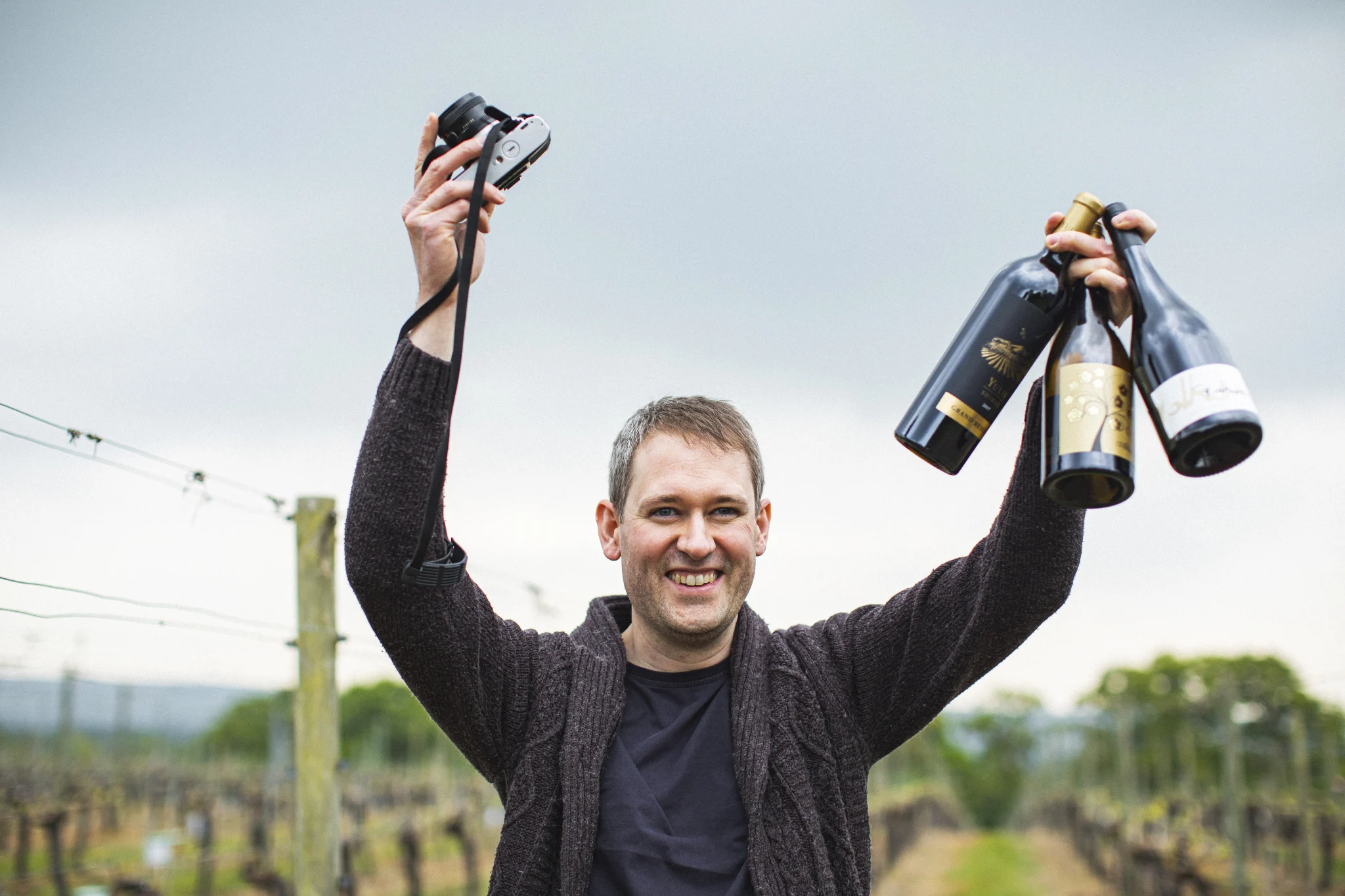 Person standing in a vineyard holding three wine bottles in one hand and a camera in the other.