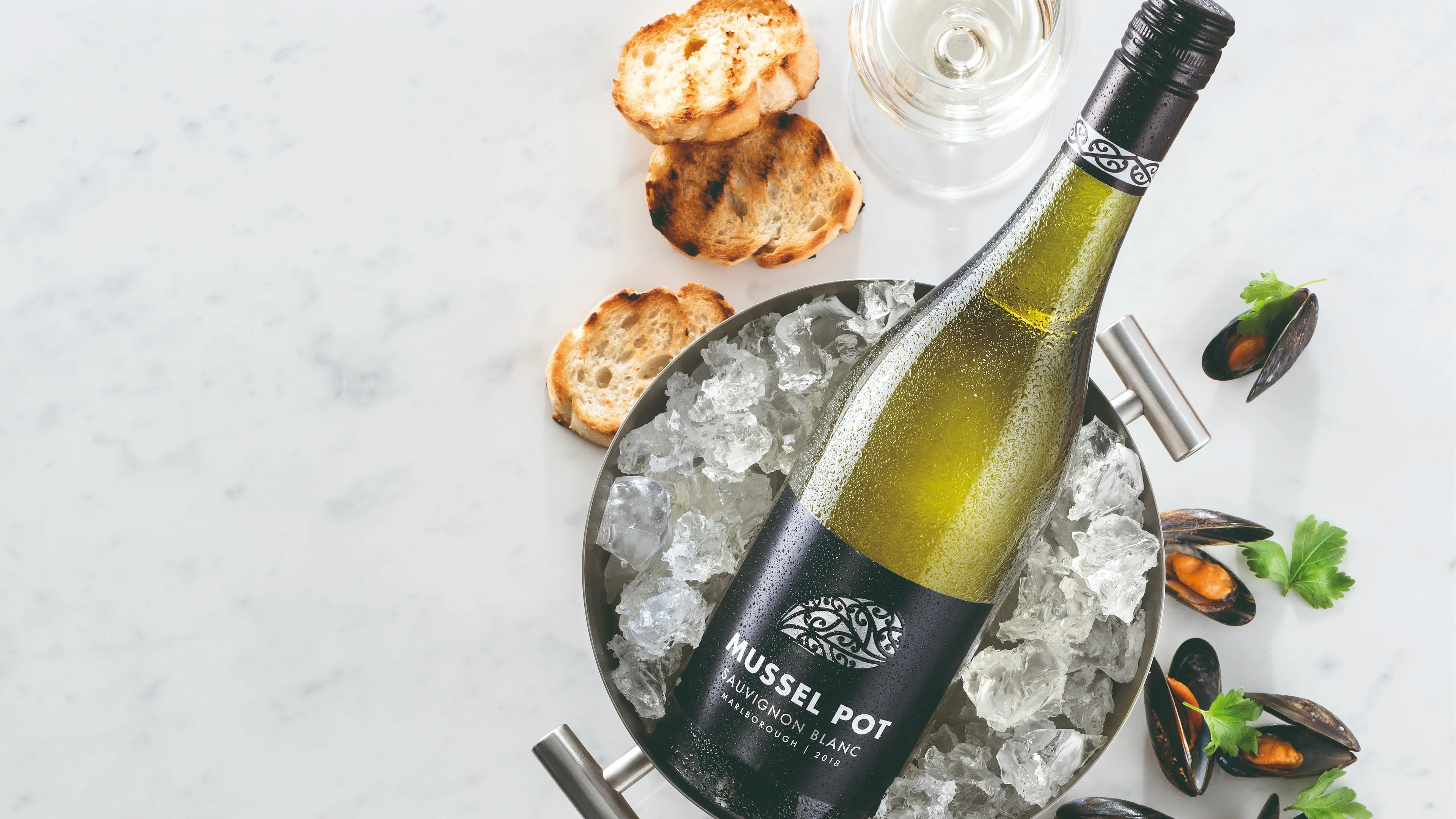 Bottle of Sauvignon Blanc chilling in an ice bucket beside a wine glass, mussels and toasted bread on a marble surface.