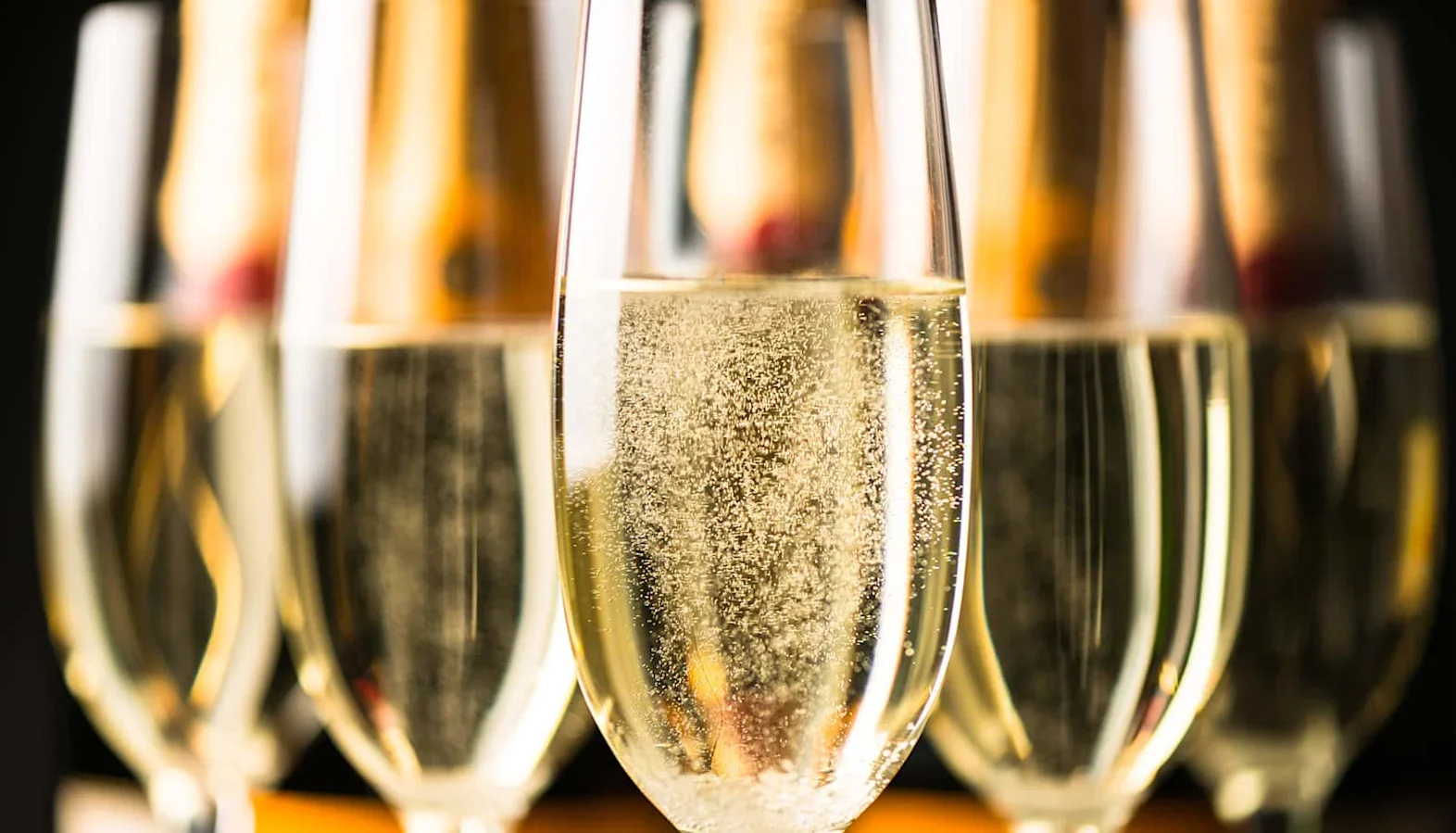 Close-up of several champagne flutes filled with sparkling wine, showing rising bubbles with blurred bottles in the background.