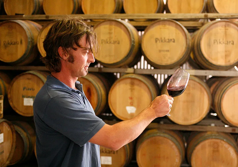 Person holding a glass of red wine in front of stacked wooden barrels inside a wine cellar.