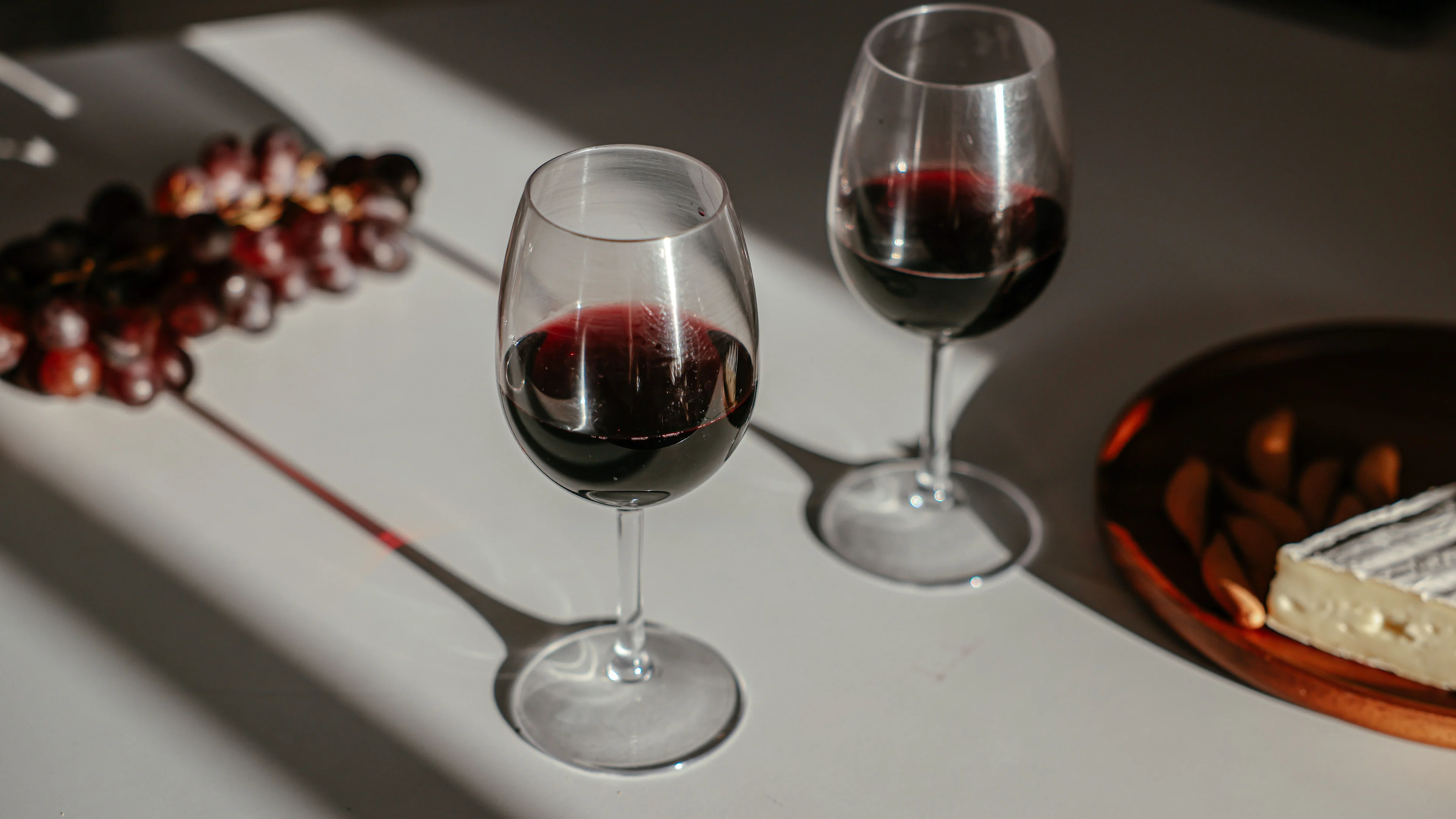 Two glasses of red wine on a table beside grapes and a cheese board in natural sunlight.