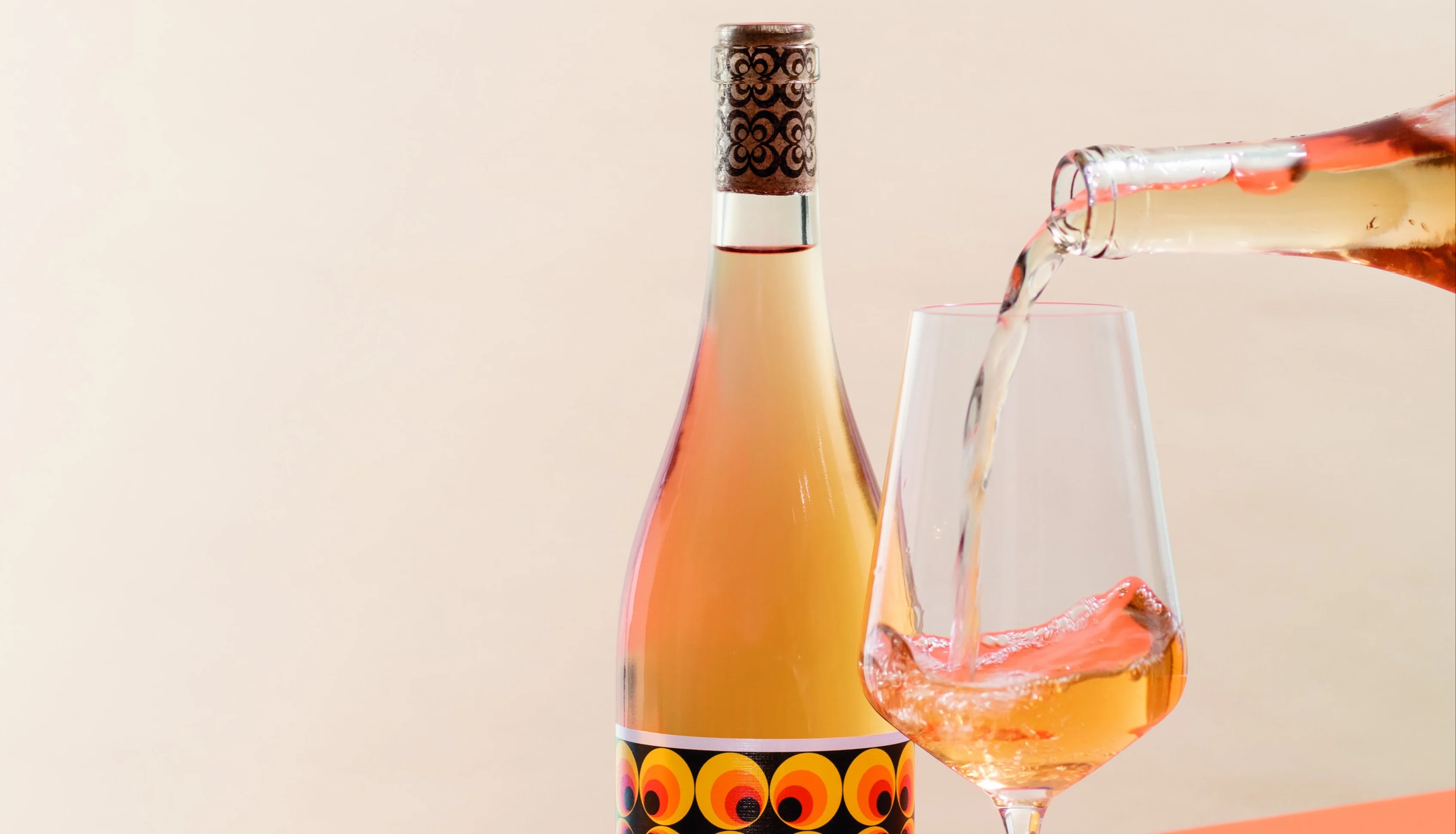 Bottle of orange wine beside a filled glass as more orange wine is poured from a second bottle against a soft peach background.