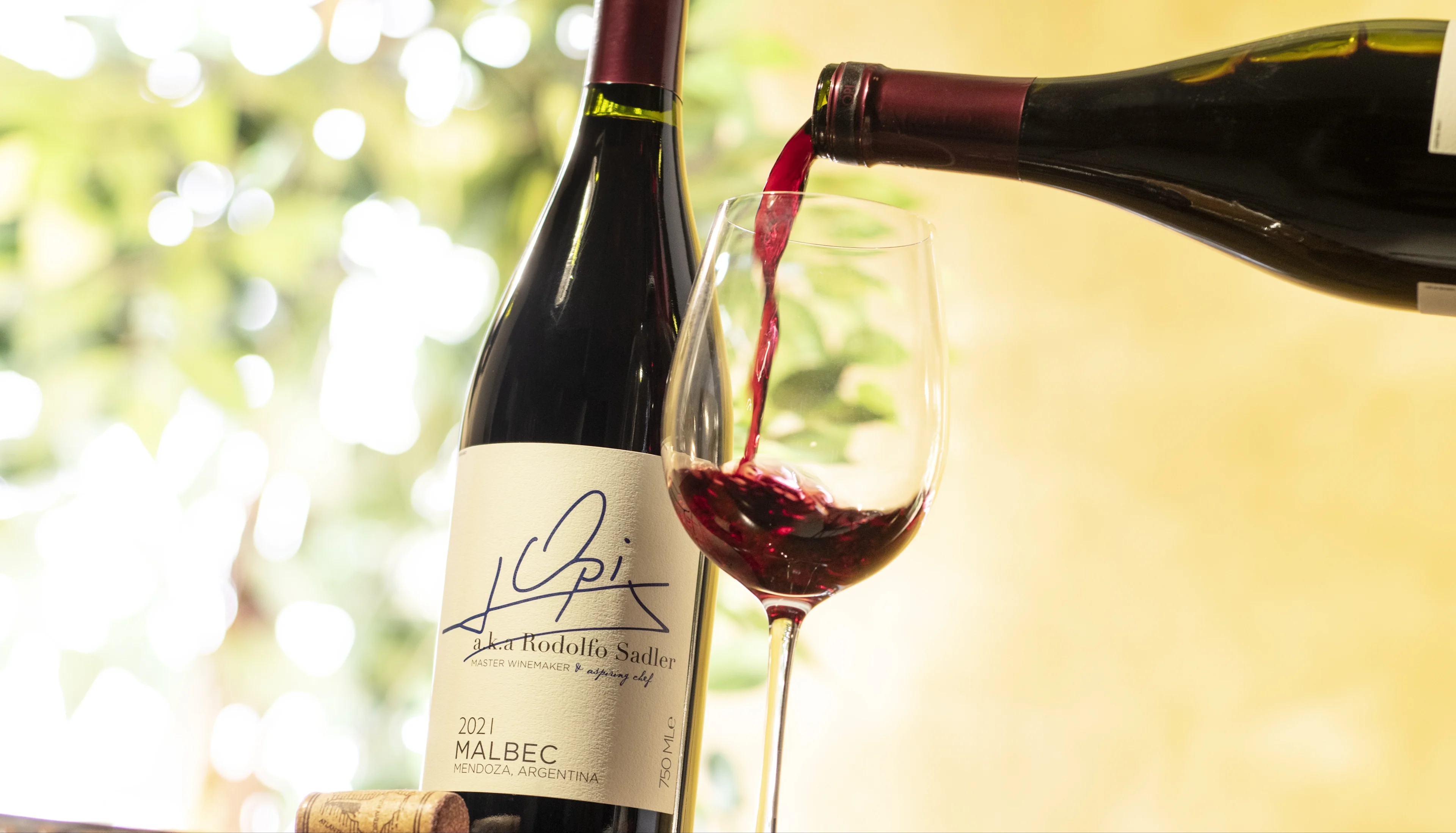 Red wine being poured into a glass beside a Malbec bottle on a wooden surface.