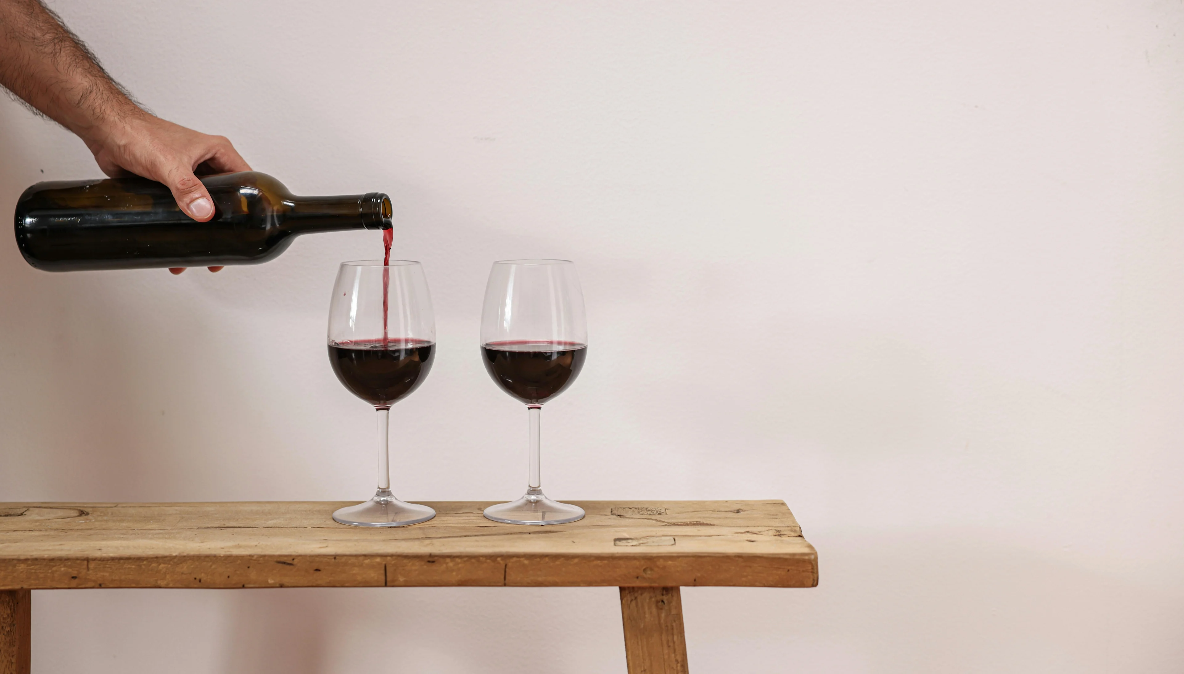 Red wine being poured into a glass beside another filled glass on a wooden bench against a plain wall.