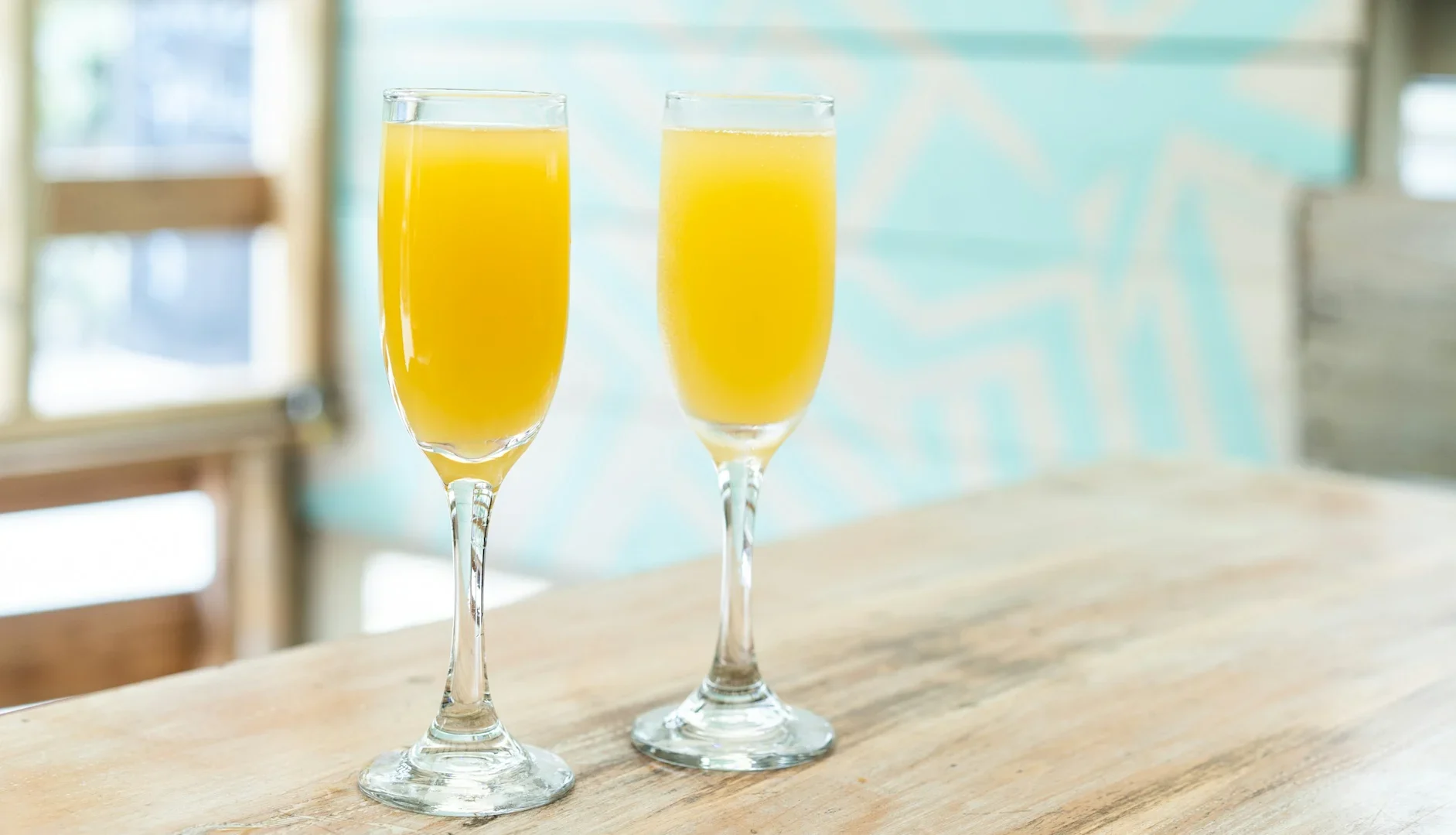 Two mimosa cocktails in tall flutes on a light wooden table.