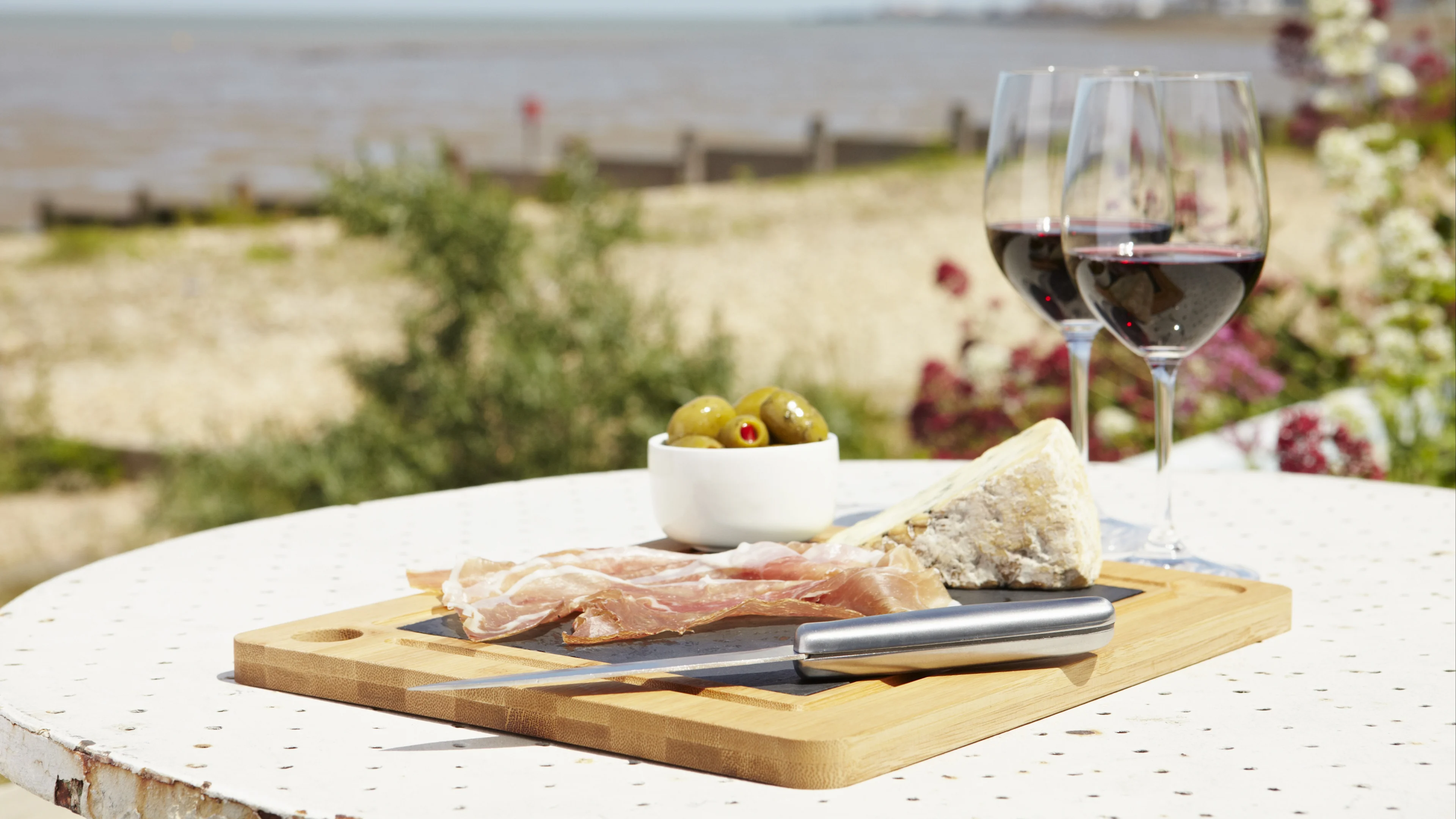 Charcuterie board with cured meat, cheese and olives served with two glasses of red wine by the seaside.