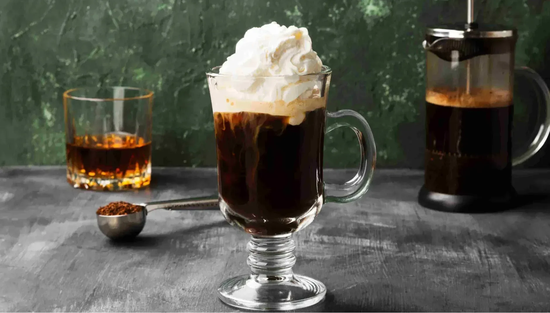 Glass mug of Irish coffee topped with whipped cream beside a French press and a glass of whiskey.