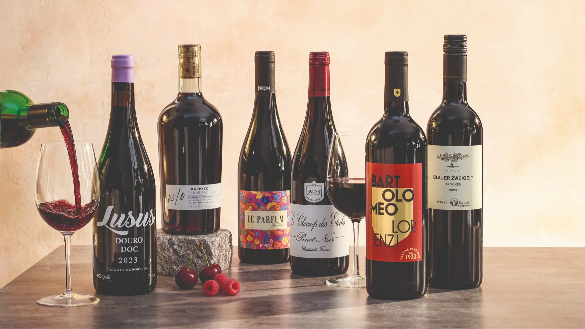 Assortment of red wine bottles with wine being poured into a glass, with raspberries and cherries on the table alongside the bottles.