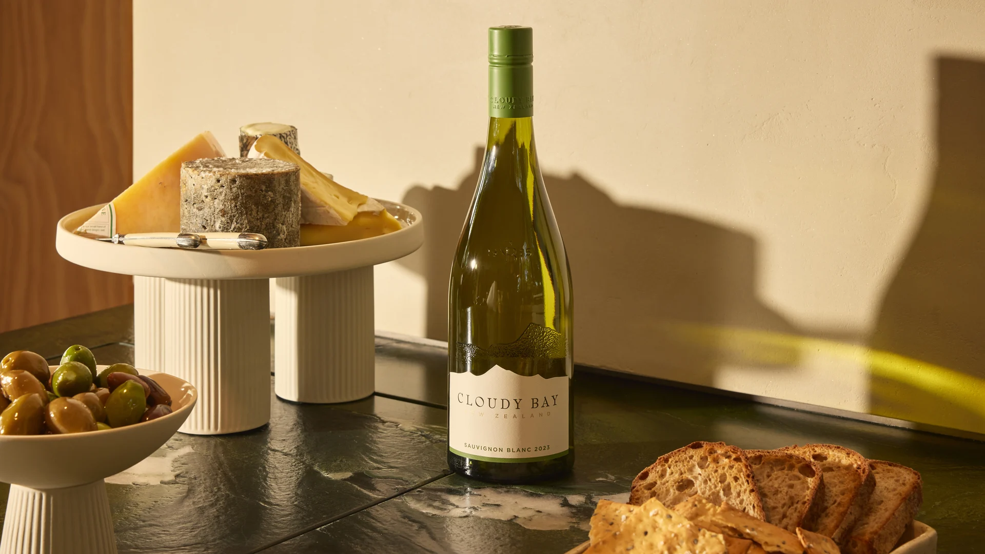 Bottle of Sauvignon Blanc surrounded by cheeses, olives and sliced bread on a kitchen counter in warm afternoon light.