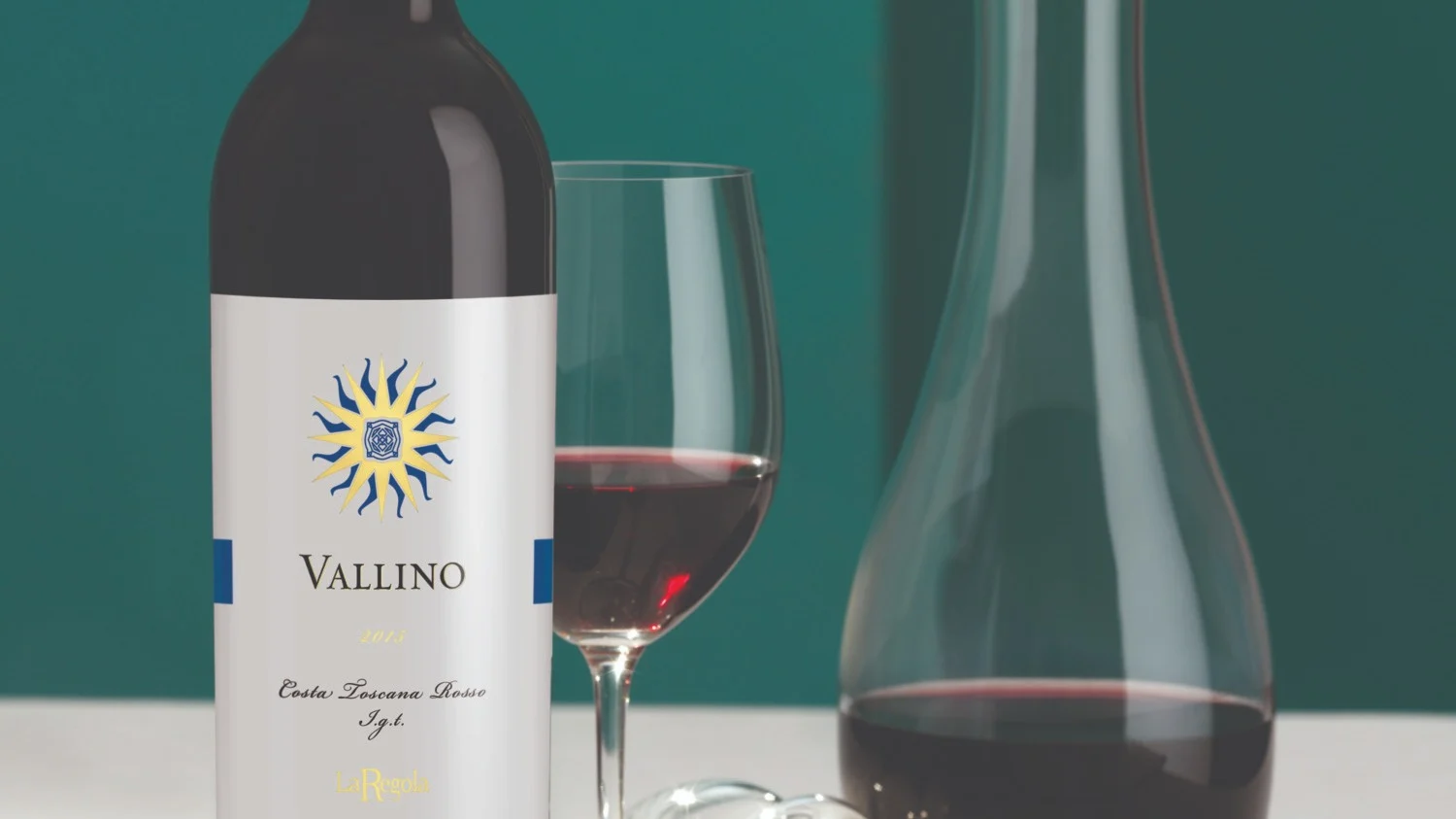Bottle of Vallino red wine with a filled wine glass, glass decanter and overturned glass on a white surface against a green background.