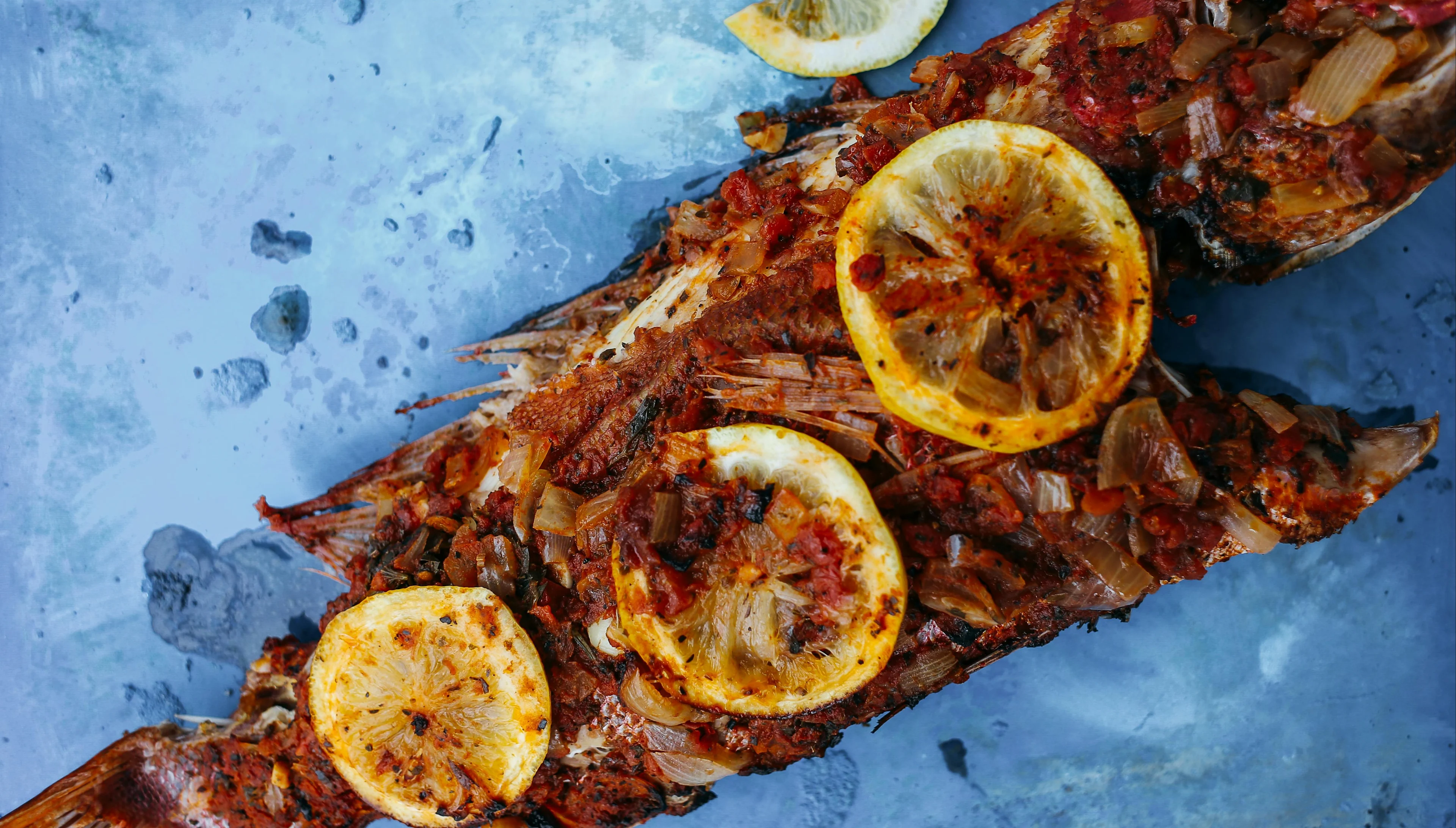 Grilled whole fish topped with lemon slices, herbs, and spices on a blue surface.
