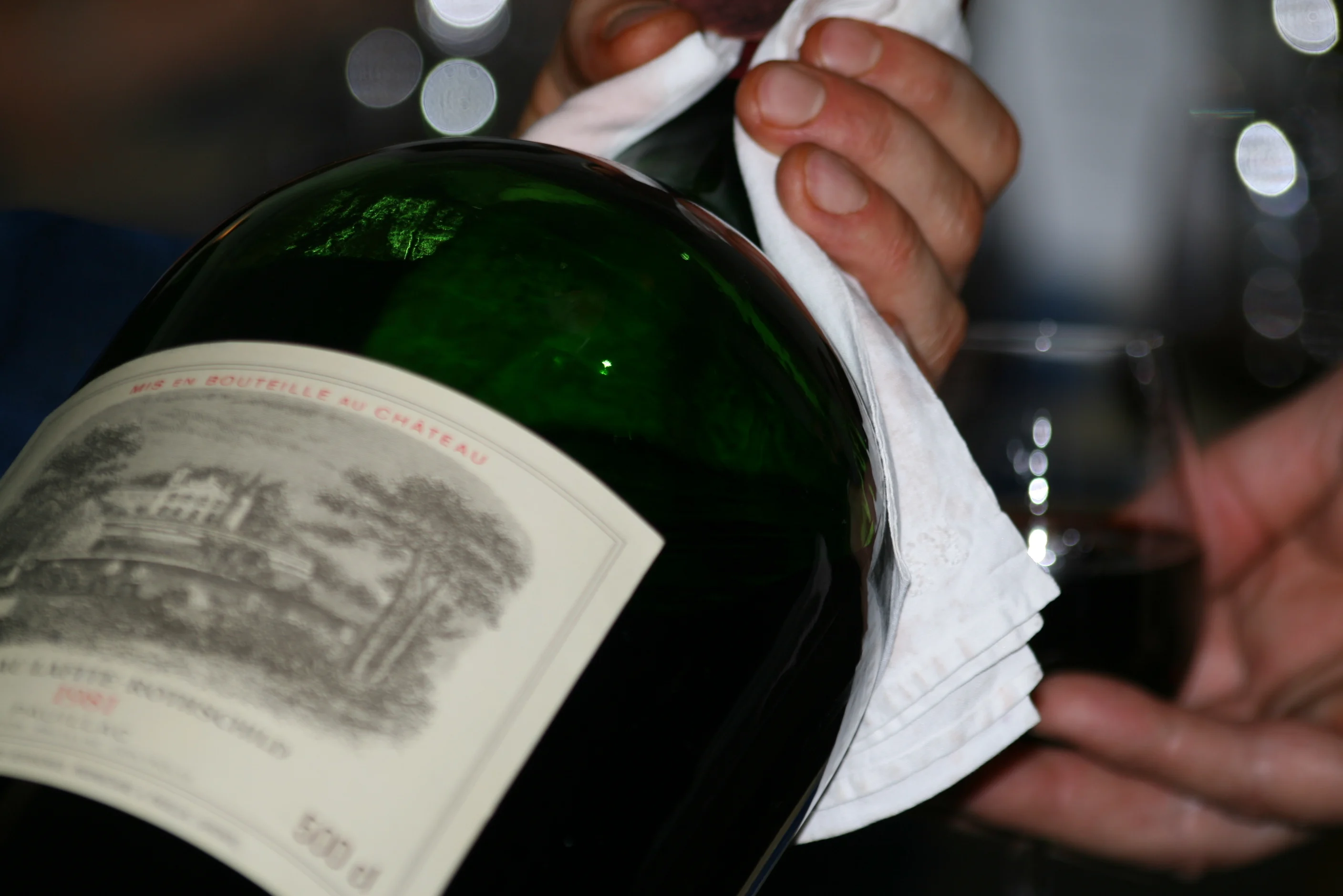 Hands polishing a large green wine bottle containing red wine with a cloth, with a glass of red wine in the background.