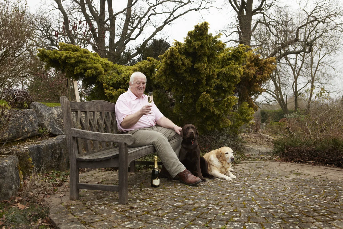 Person seated on a garden bench in East Sussex holding a glass of sparkling wine with two dogs and a bottle on cobblestone paving.