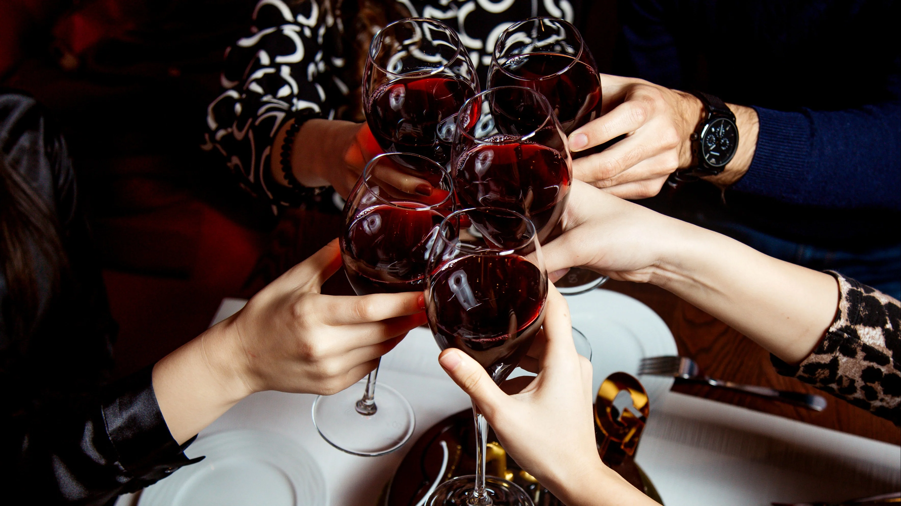 Several hands clinking glasses of red wine over a dining table set with dessert plates.