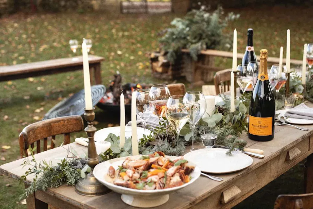Outdoor table set with champagne, glasses, candles, and sharing dishes surrounded by autumn foliage.