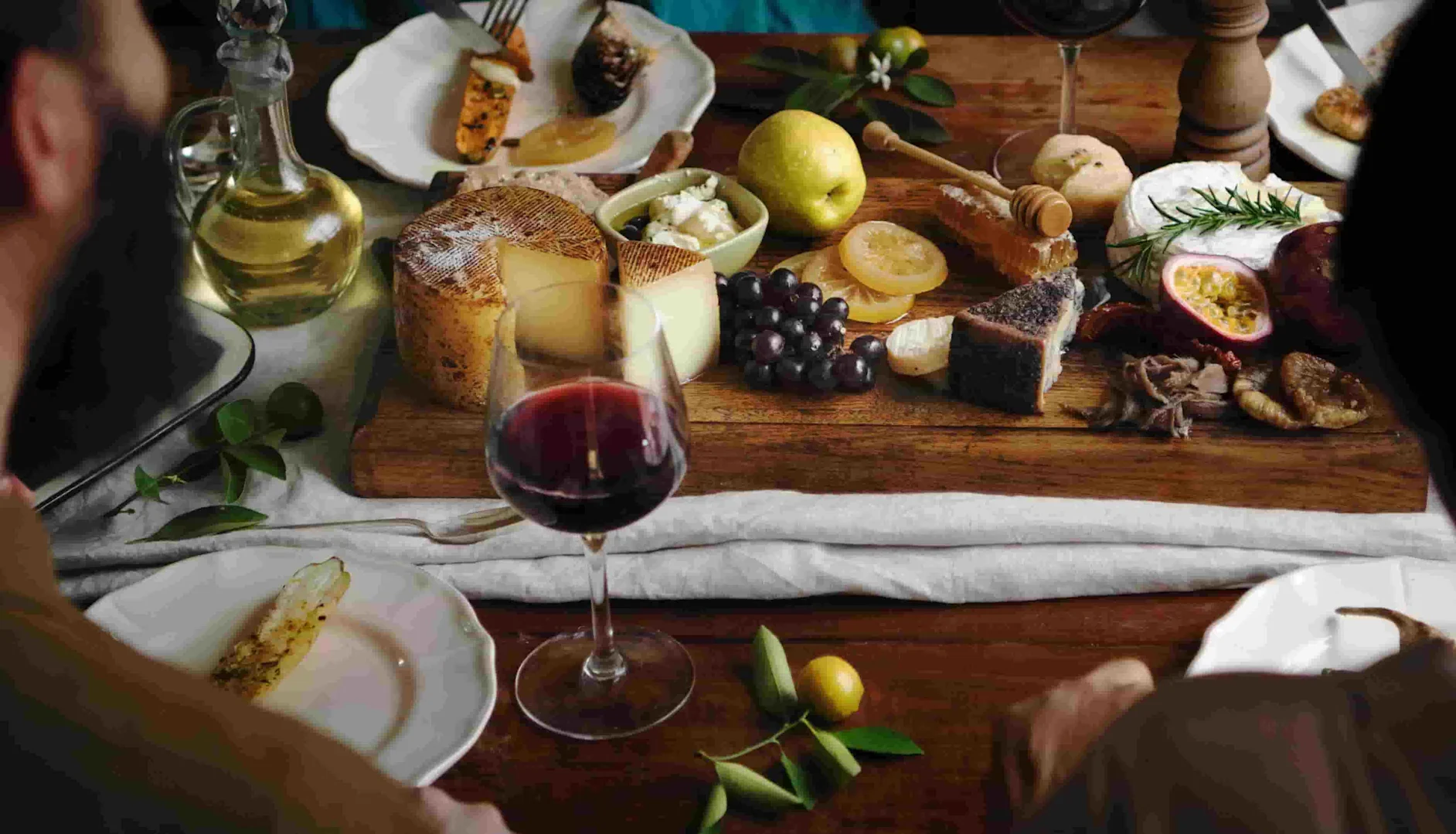 A wooden board with assorted cheeses, grapes, honeycomb, sliced fruit, and bread, surrounded by plates and glasses of red wine on a rustic dining table.