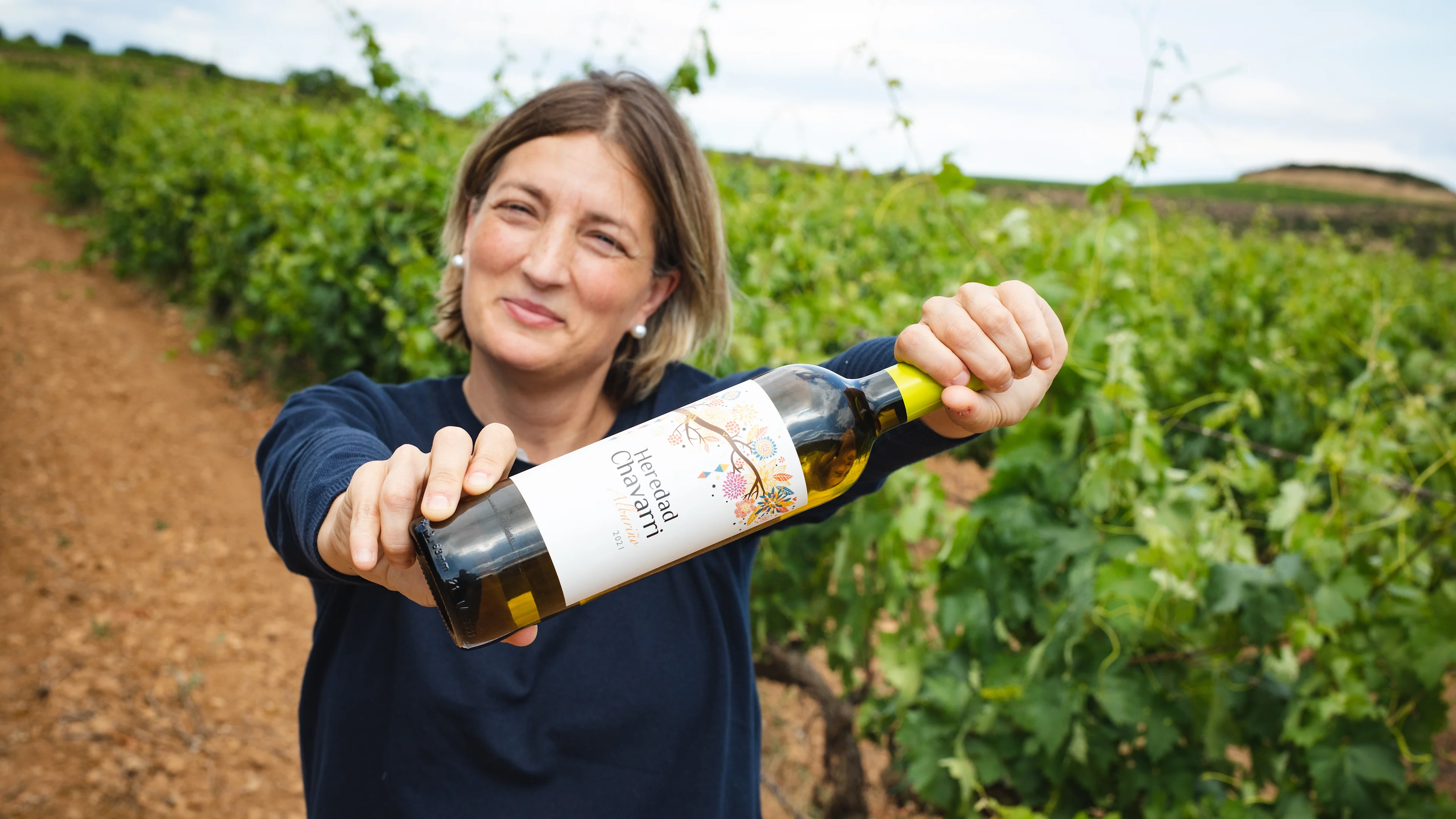 Person holding a bottle of Albariño wine while standing between vineyard rows.