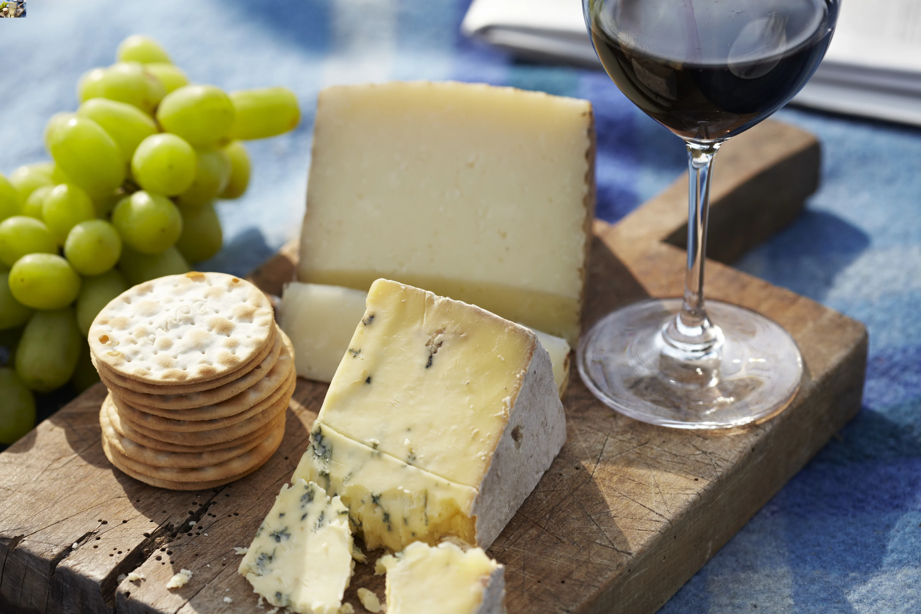A wooden board with wedges of blue and hard cheese, stacked crackers, green grapes, and a glass of red wine on a blue cloth background.